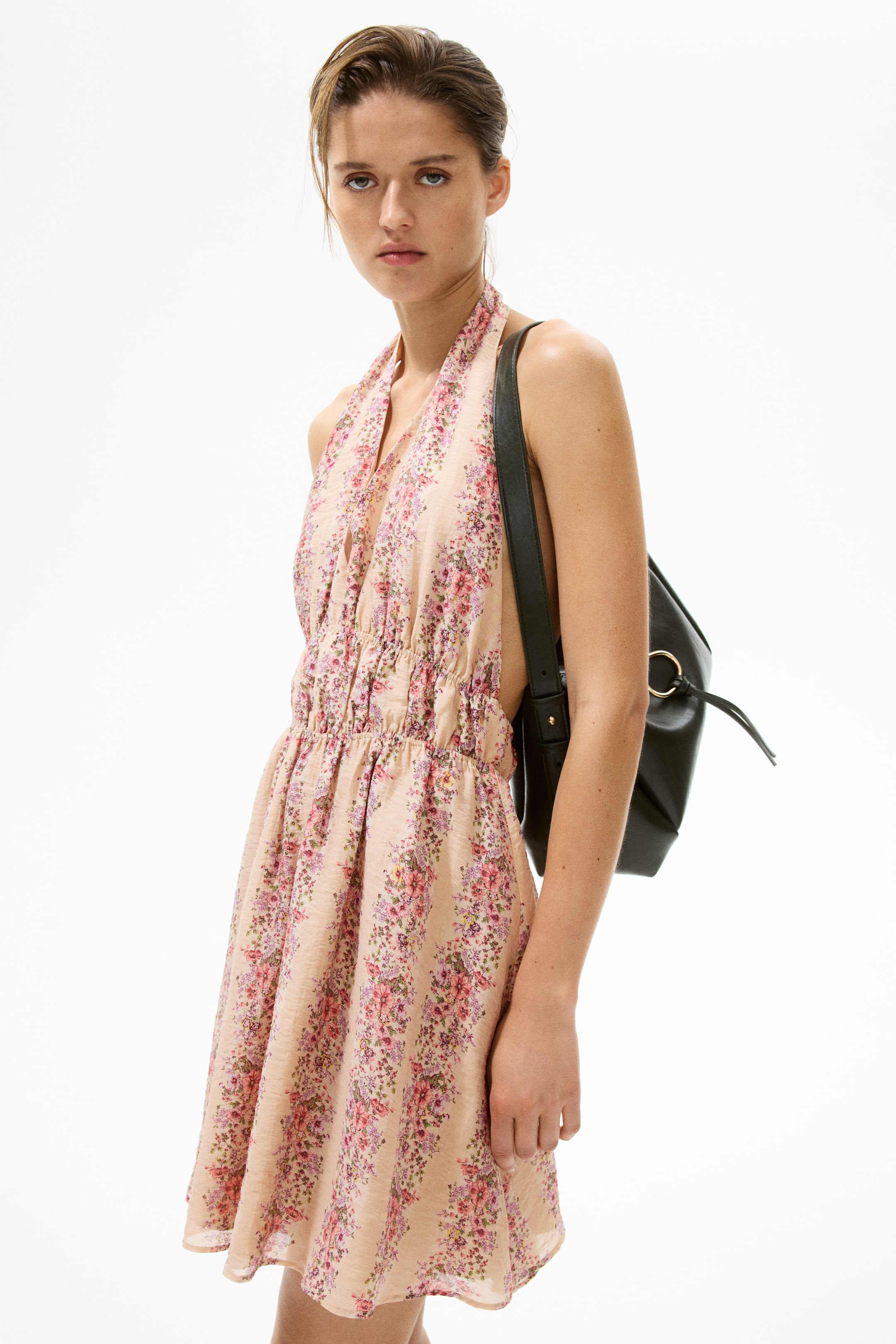 View larger image: The model is wearing a short halterneck dress adorned with a floral pattern in shades of pink and purple against a light backdrop. This dress boasts a deep V neckline, a fitted bodice with two rows of narrow elasticated detailing at the waist, and a flared skirt. The model complements the outfit with a black rucksack slung over the dress.