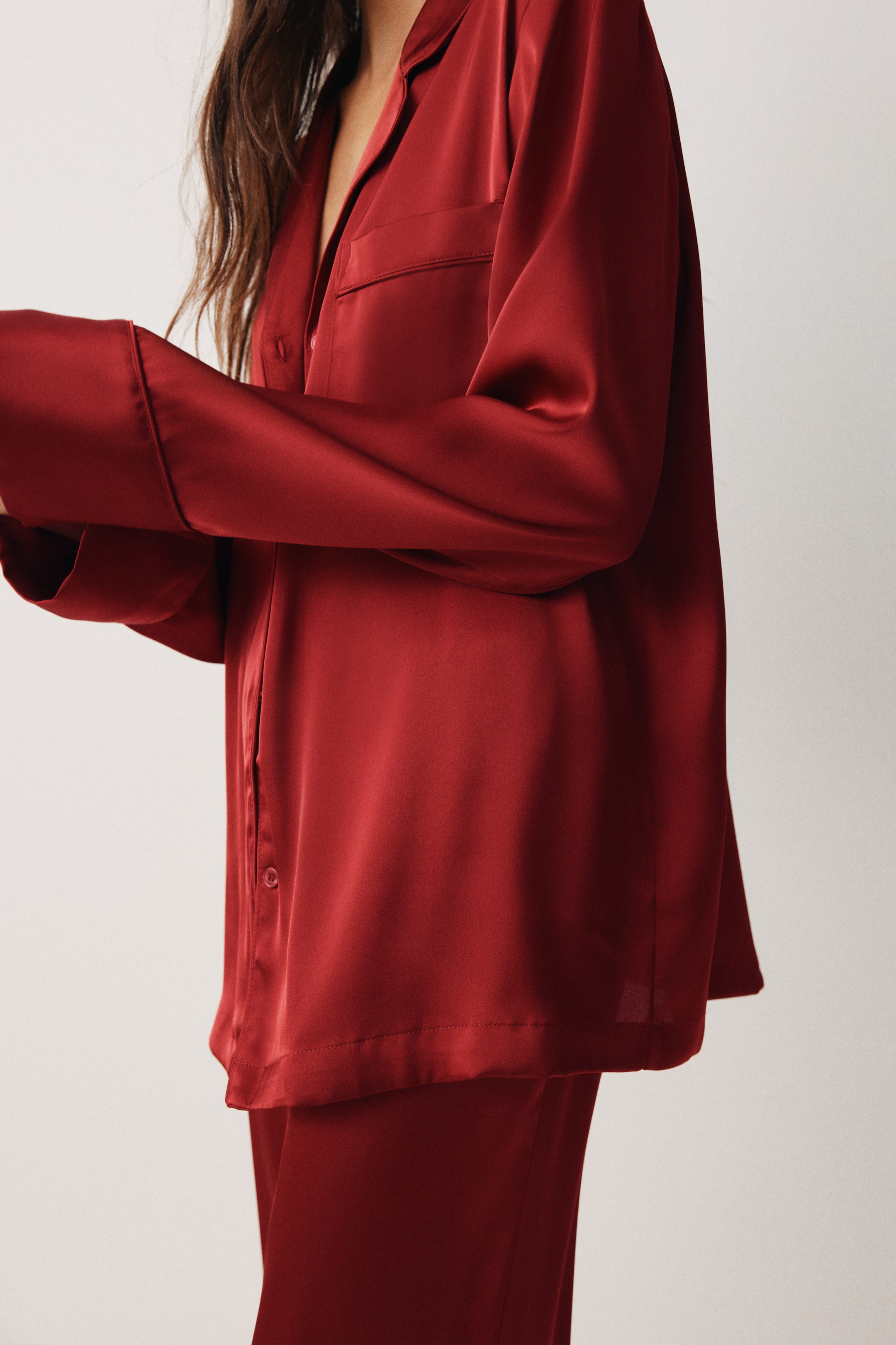 View larger image: A red satin pajama top is being presented from a slightly angled side view, featuring a notch lapel collar, a button-front closure, and a chest patch pocket. The long-sleeved top is flowing over matching red satin pants.