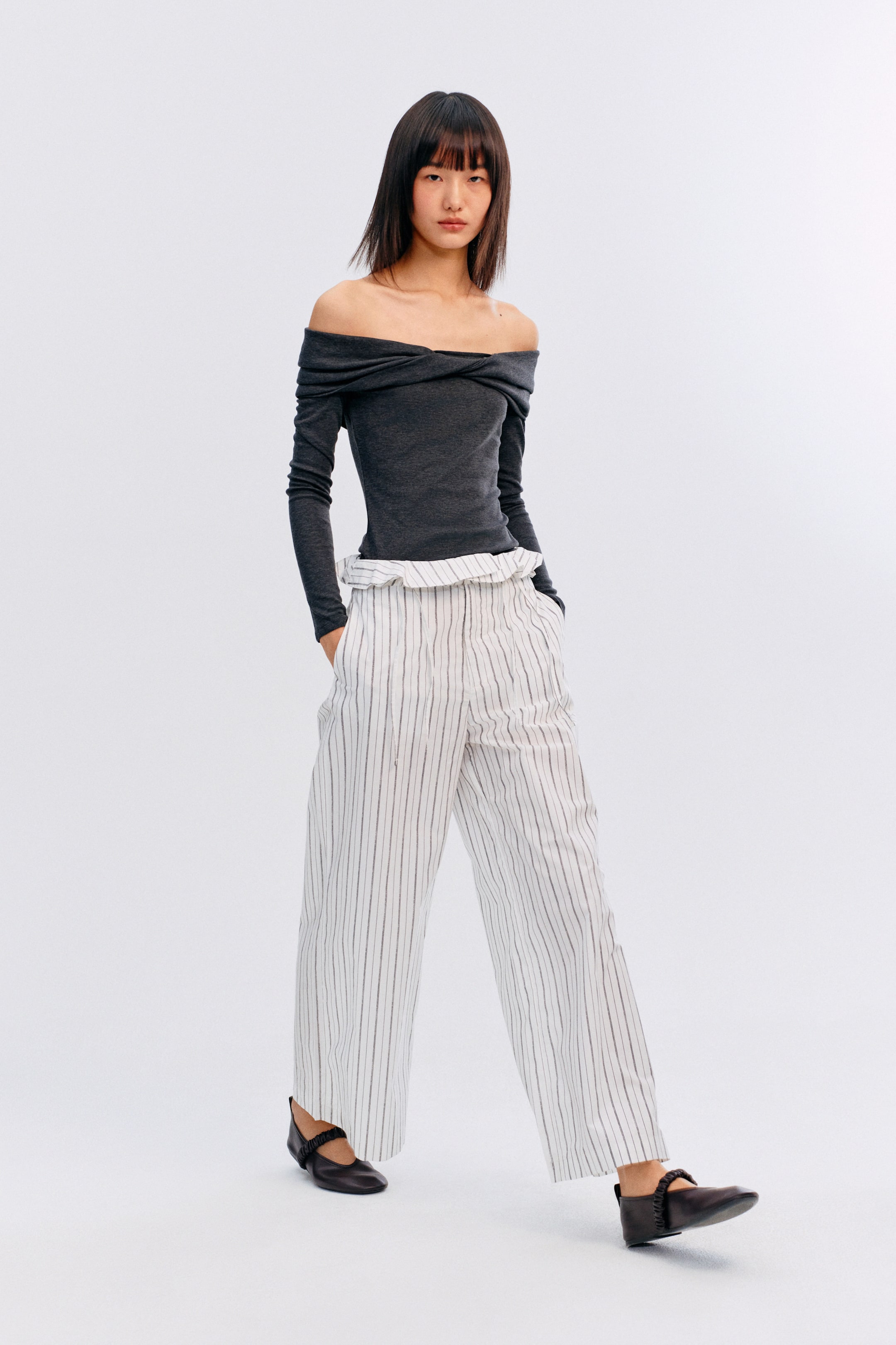 이미지 확대 보기: 어깨를 감싸는 어두운 회색 오프숄더 긴소매 탑은 흰색 와이드 레그 팬츠에 넣어 입고 있습니다. 이 팬츠는 세로 핀스트라이프, 주름이 있는 하이웨이스트, 눈에 띄는 사이드 포켓이 특징이며, 다크 브라운 메리 제인 플랫 슈즈는 발등에 주름이 있는 스트랩으로 전체적인 스타일을 완성합니다.