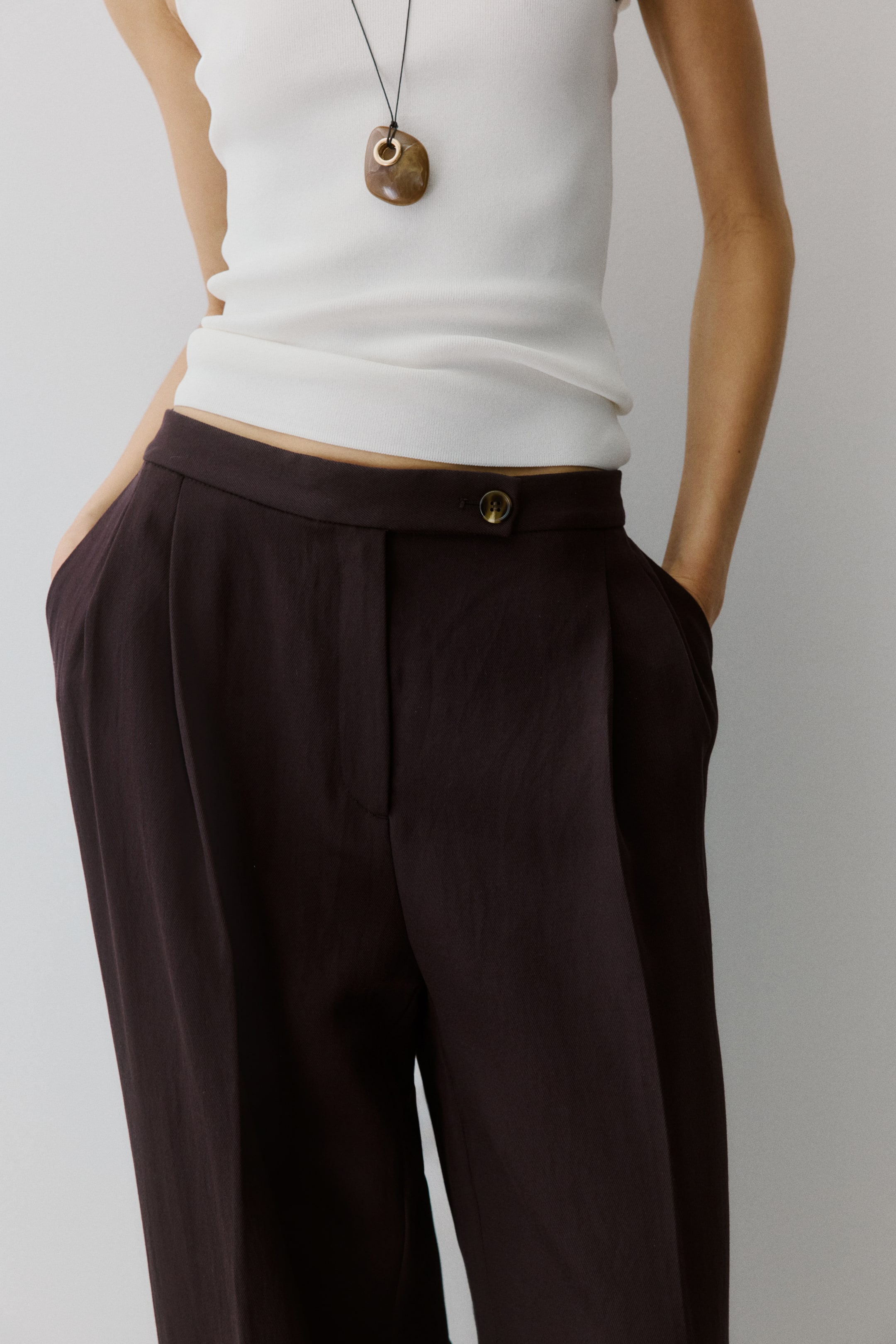 Voir une image plus grande: Le mannequin porte un pantalon ample de couleur brun foncé en tissu tissé, doté d'une ceinture allongée avec fermeture à bouton, d'une braguette à glissière, de jambes larges avec plis à l'avant et de plis marqués sur le devant. Le mannequin place ses mains dans les poches latérales en biais.