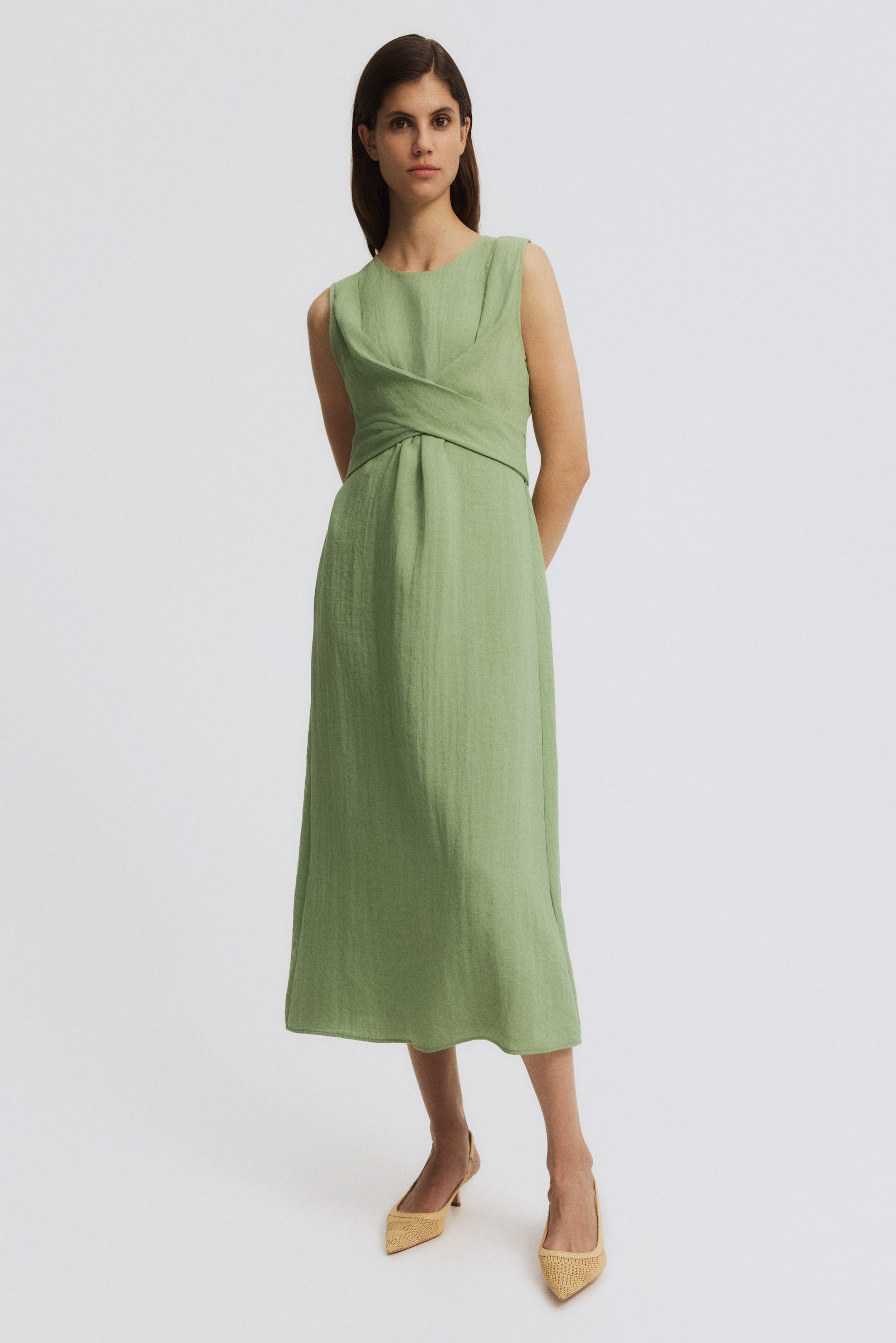 Agrandir l'image: Une robe midi verte sans manches présente un tissu texturé et un col rond. Le corsage met en avant un détail croisé à la taille, avec des fronces s'étendant dans la jupe longueur midi.