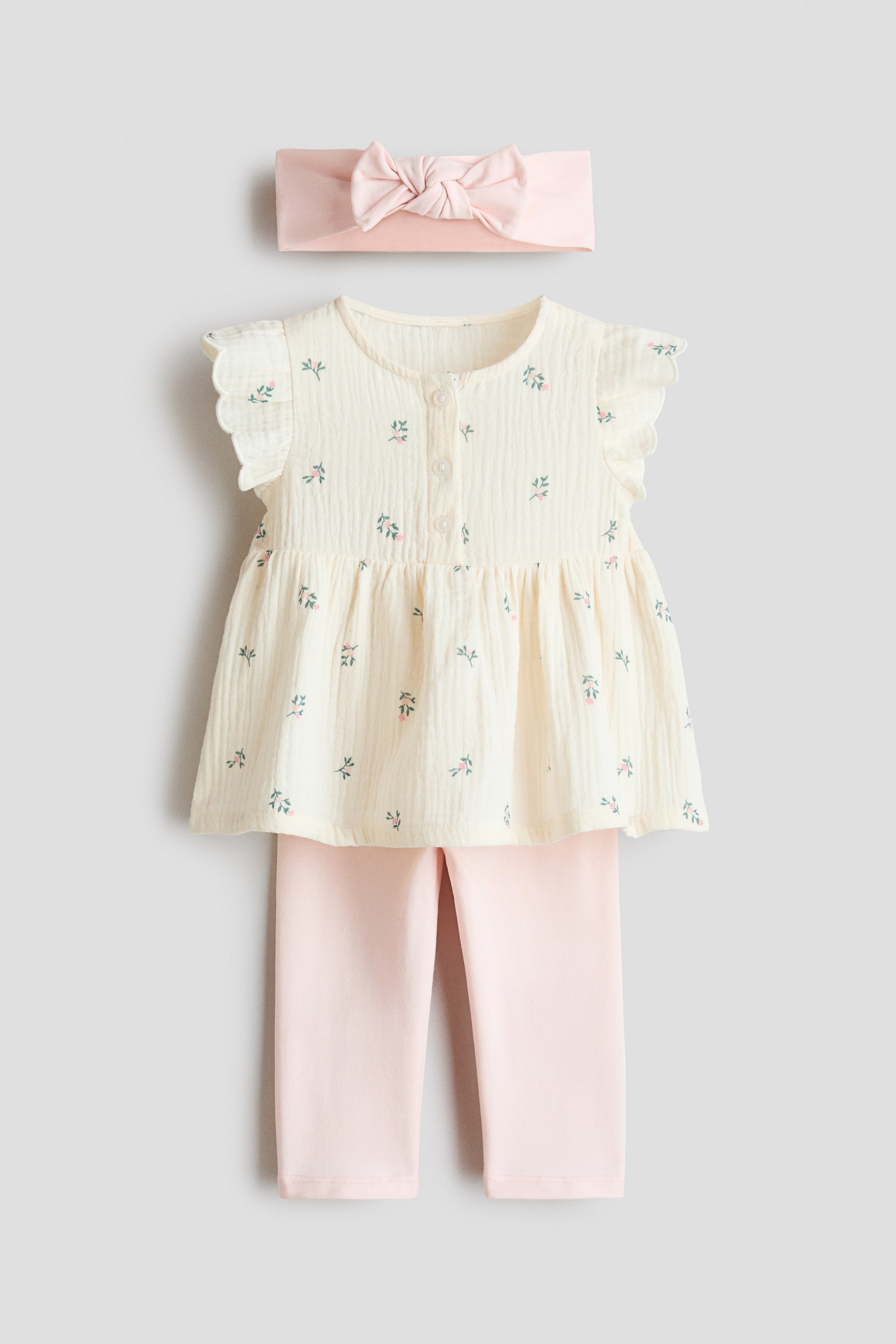 View larger image: A three-piece baby outfit showcases a cream-coloured, crinkled cotton tunic featuring a button placket, ruffled cap sleeves, and a small pink and green floral print. It is paired with plain pink leggings and a matching pink knotted headband, all facing front.