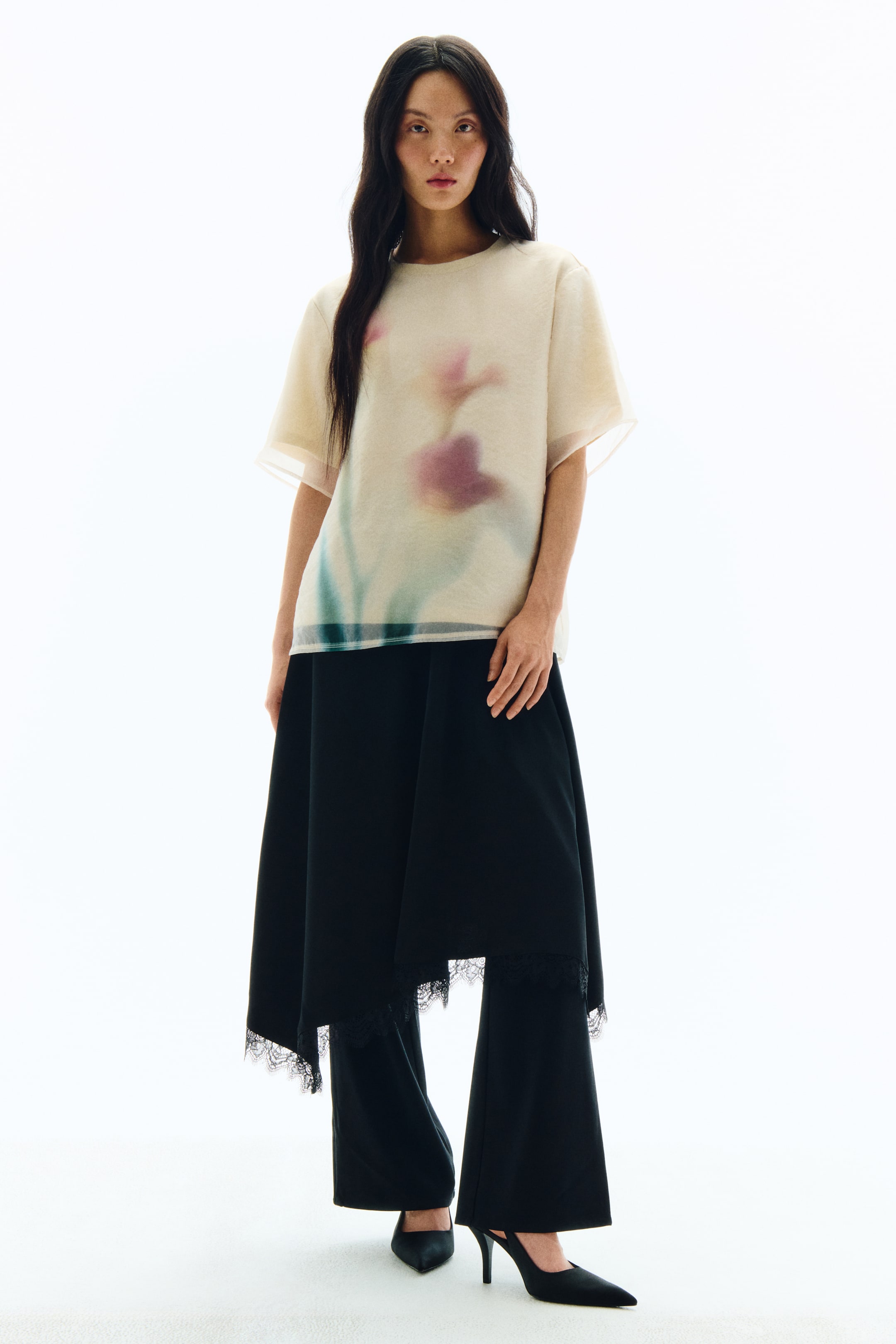 View larger image: The product is a complete outfit featuring a sheer, cream-coloured short-sleeved top with a blurred pink and teal floral print, styled over black wide-leg trousers. A black asymmetrical midi skirt with a lace-trimmed hem is layered over the trousers, and black pointed-toe slingback heels are visible.