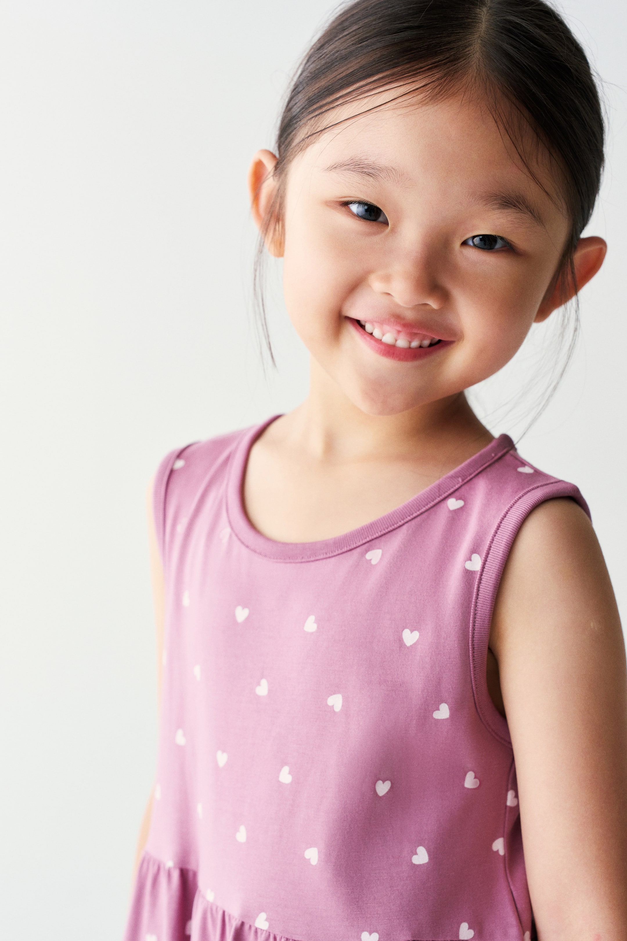View larger image: The model is wearing a mauve-pink sleeveless dress with an all-over white heart pattern. It features a round neckline with ribbed trim and a gathered waistline.