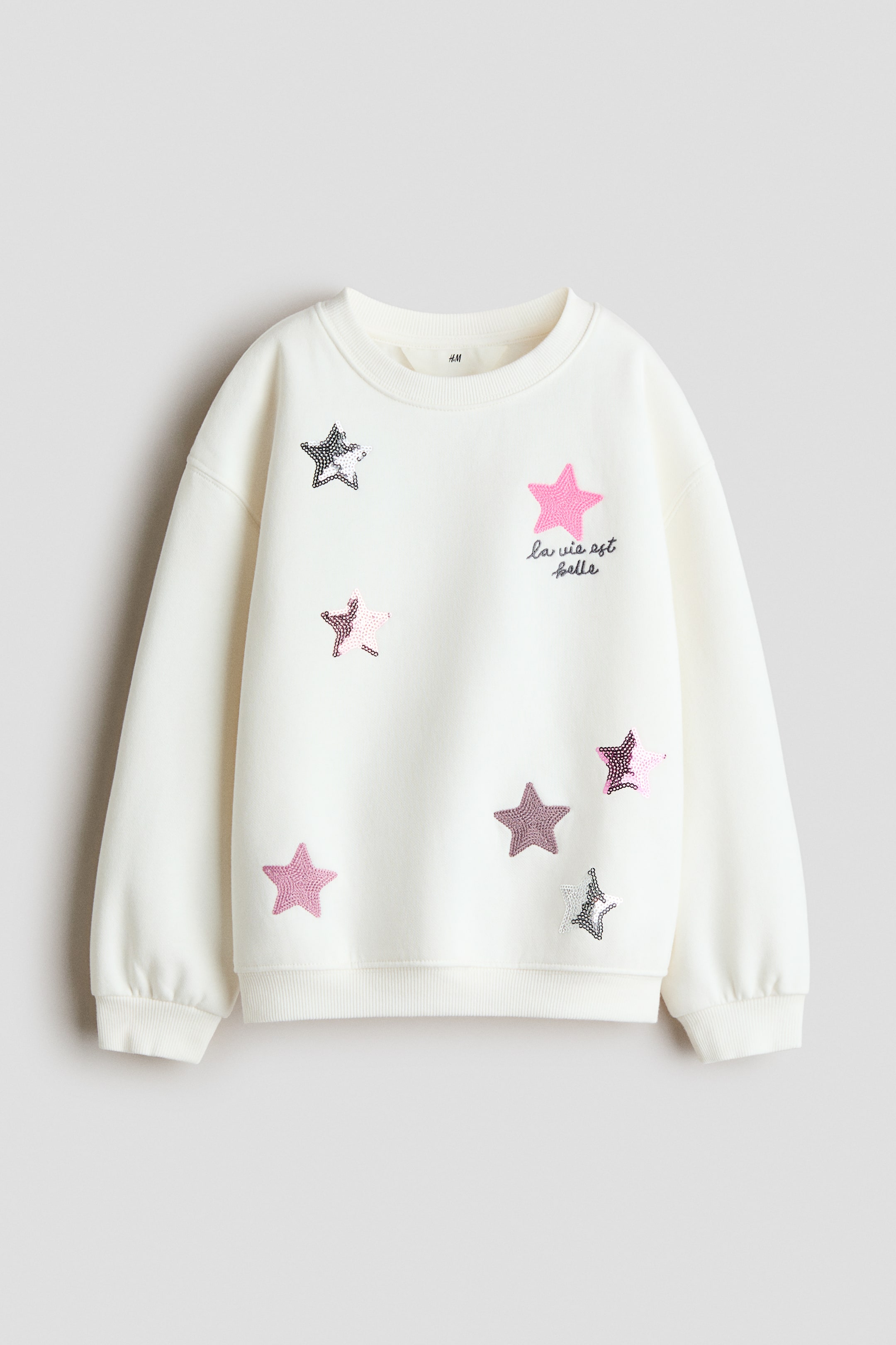View larger image: A cream sweatshirt is shown from the front, featuring a rib-trimmed crew neckline, dropped shoulders, and ribbed cuffs and hem. The front is decorated with multiple sequined stars in shades of pink, silver, and black, along with the embroidered text "la vie est belle" in black script.