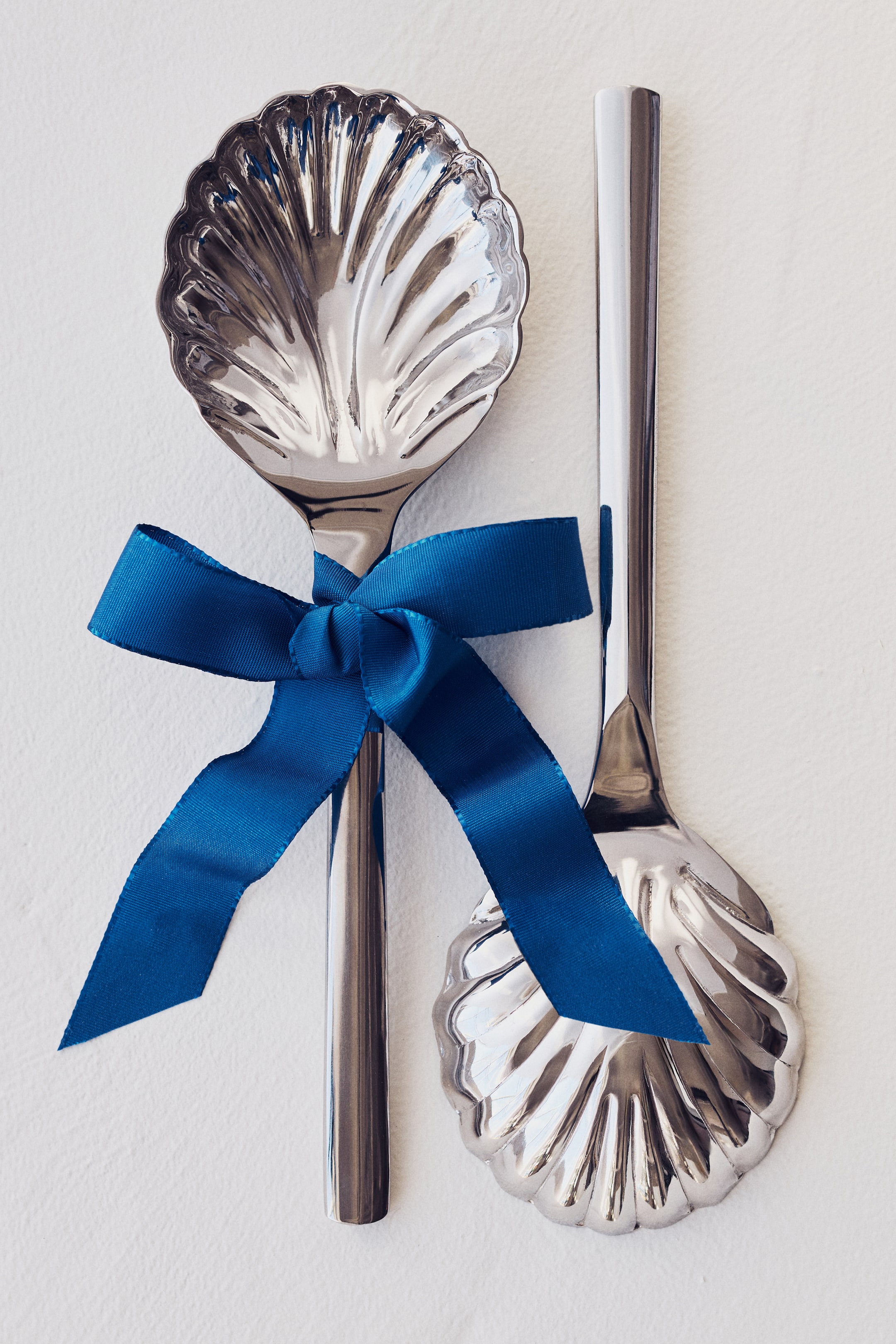 View larger image: Salad servers exhibit a polished stainless steel finish, with their heads intricately shaped like seashells. A vibrant blue ribbon ties the two servers together around their handles, presenting them from the front.