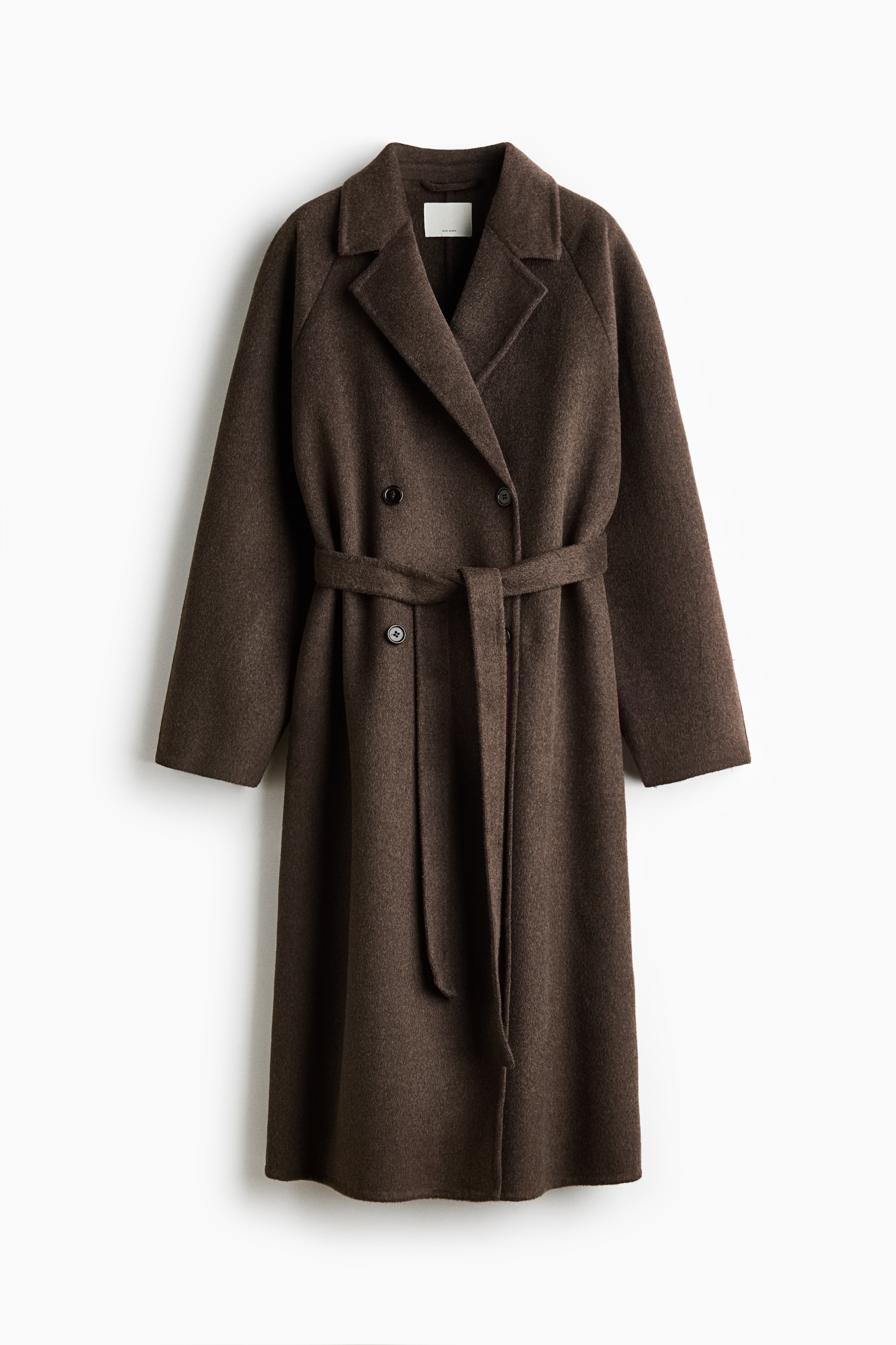 A long brown wool-blend coat featuring notch lapels, raglan sleeves, and double-breasted button fasteners is worn by the model over a black turtleneck. The model is holding a structured black handbag with gold-toned hardware under one arm.