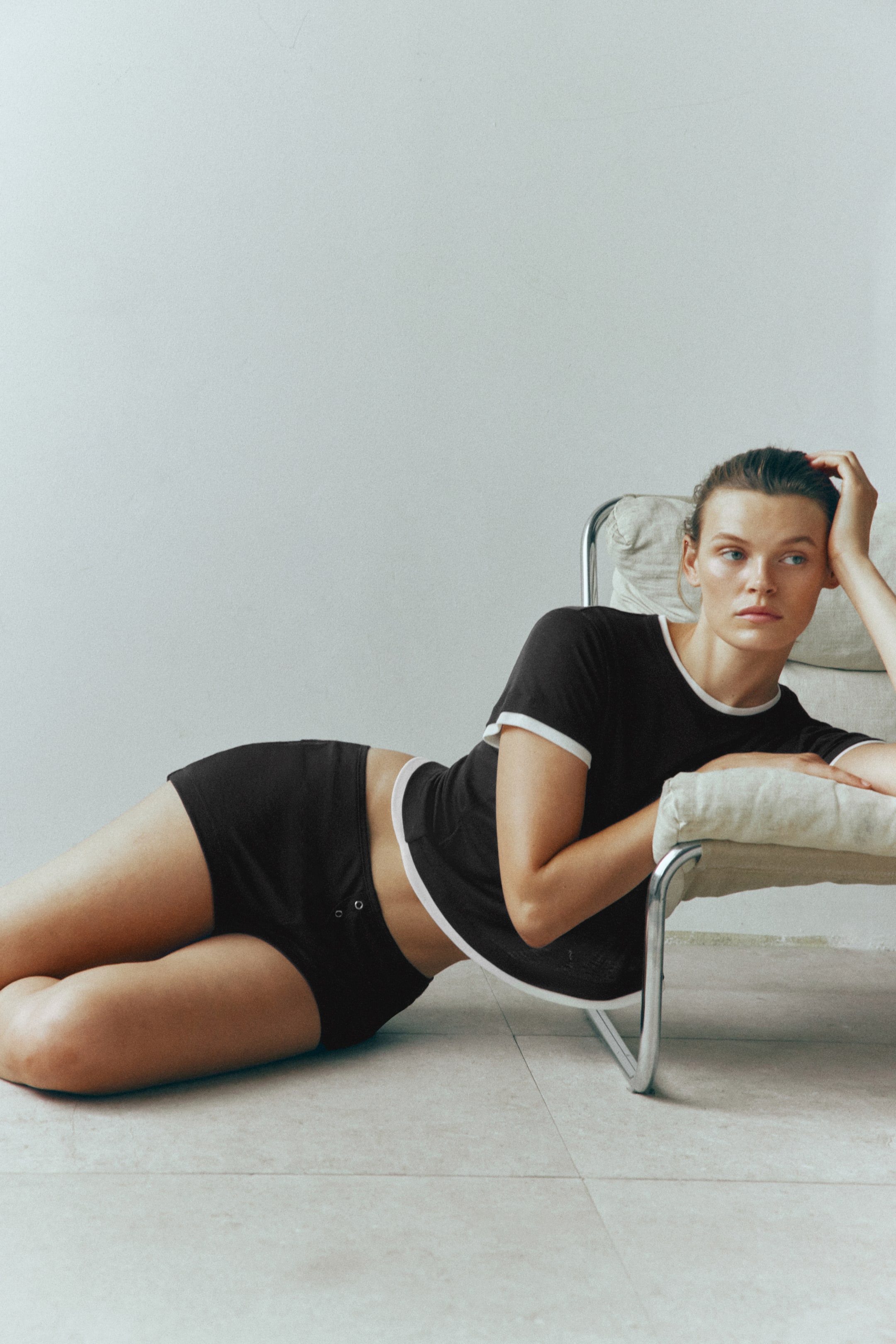 View larger image: A black ribbed jersey pyjama set is being worn by the model reclining on the floor. The set consists of a short-sleeved top with a round neckline and white contrast borders, and matching shorts featuring a mock fly with decorative press studs.