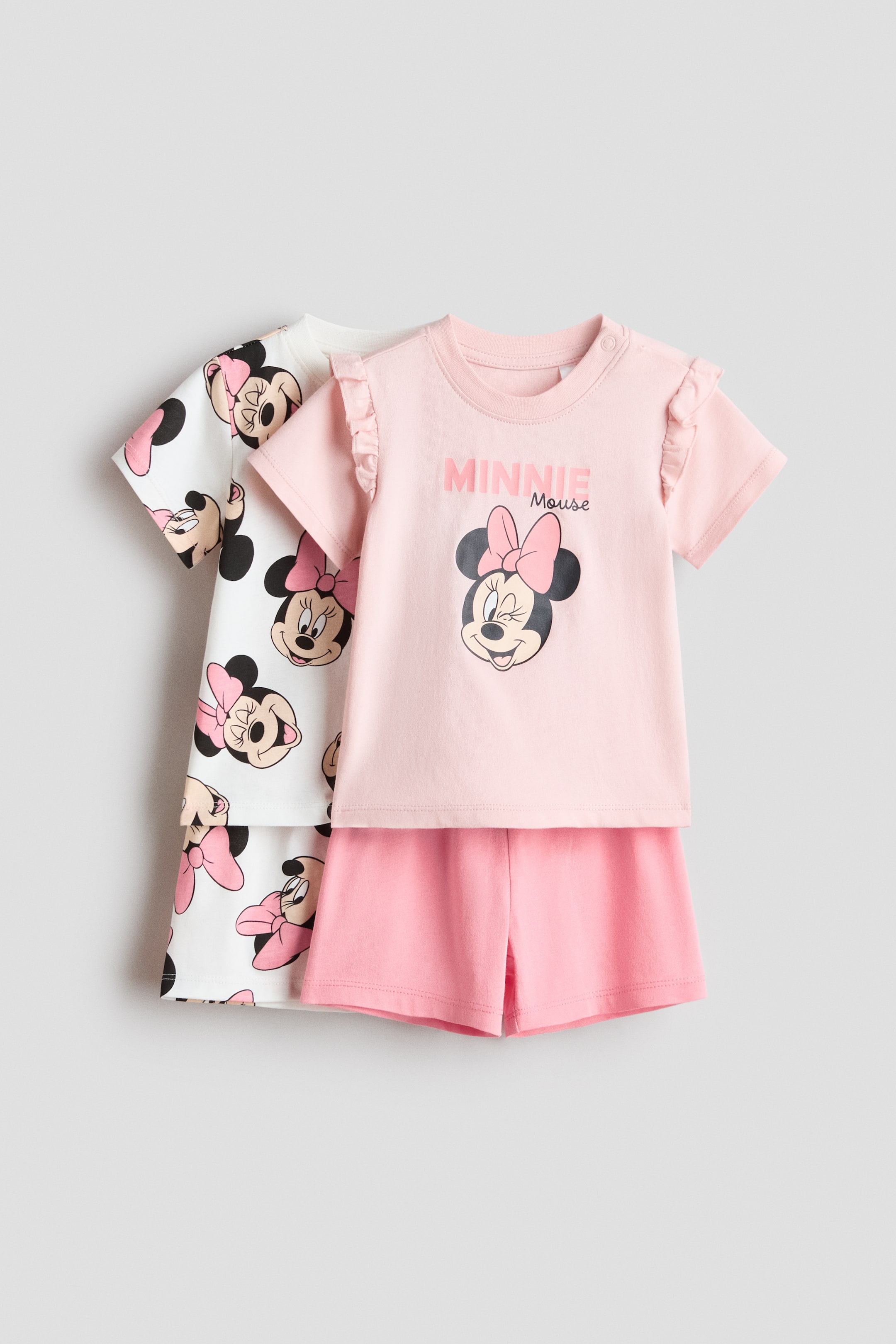 Agrandir l'image: Lot de 2 pyjamas imprimés. Le premier ensemble présente un haut à manches courtes rose clair avec un col rond, des boutons-pression sur une épaule, des manches à volants et un imprimé graphique Minnie Mouse, assorti à un short rose uni. Le deuxième ensemble, placé derrière, montre un haut à manches courtes blanc et un short assorti, tous deux recouverts d'un imprimé répété de têtes de Minnie Mouse.