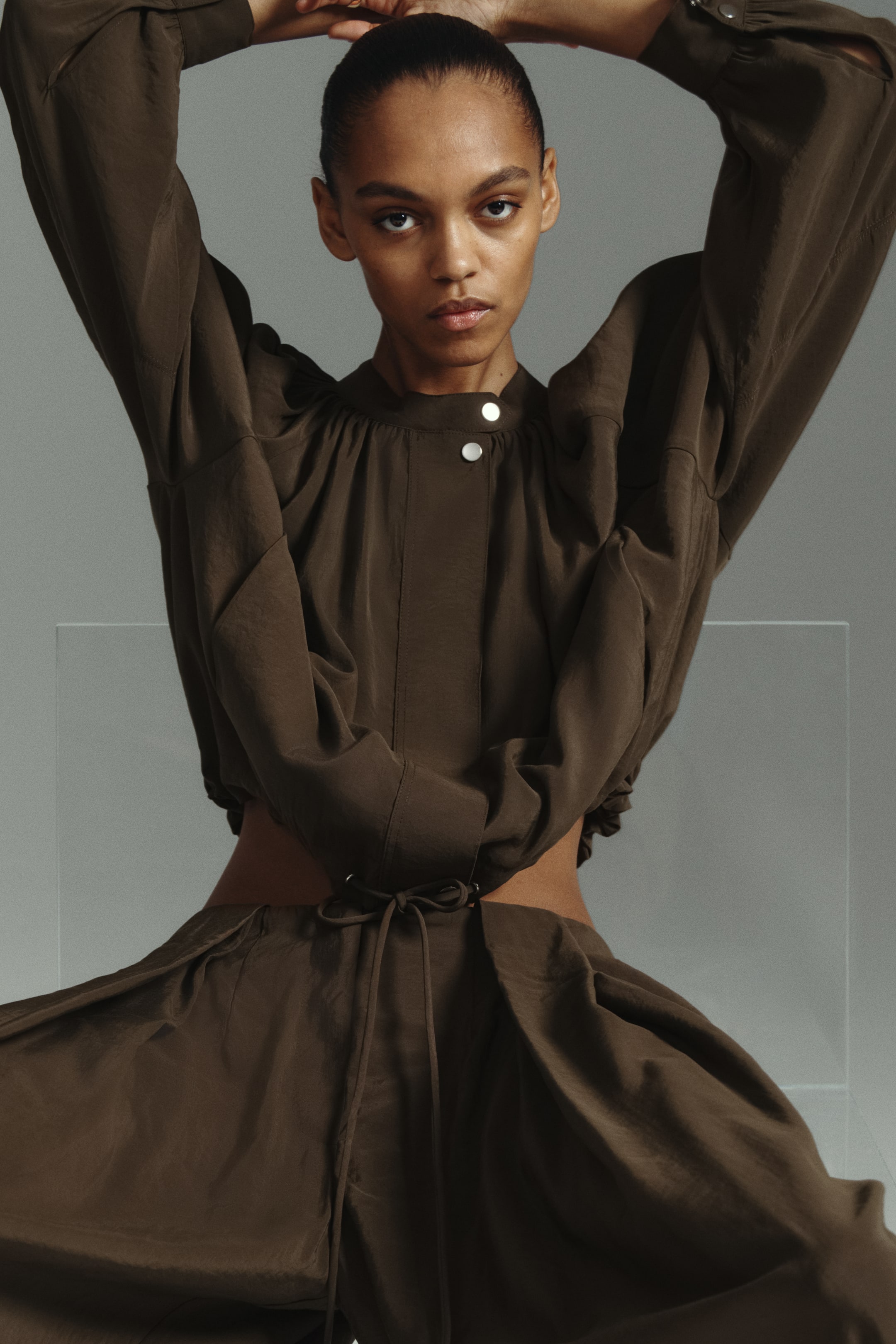View larger image: The model is showcasing an oversized jacket crafted from a dark, softly draping fabric. It features a gathered band collar, a front placket with two visible buttons, and long balloon sleeves with press-studs at the cuffs. The drawstring hem is tied, accentuating the waist.