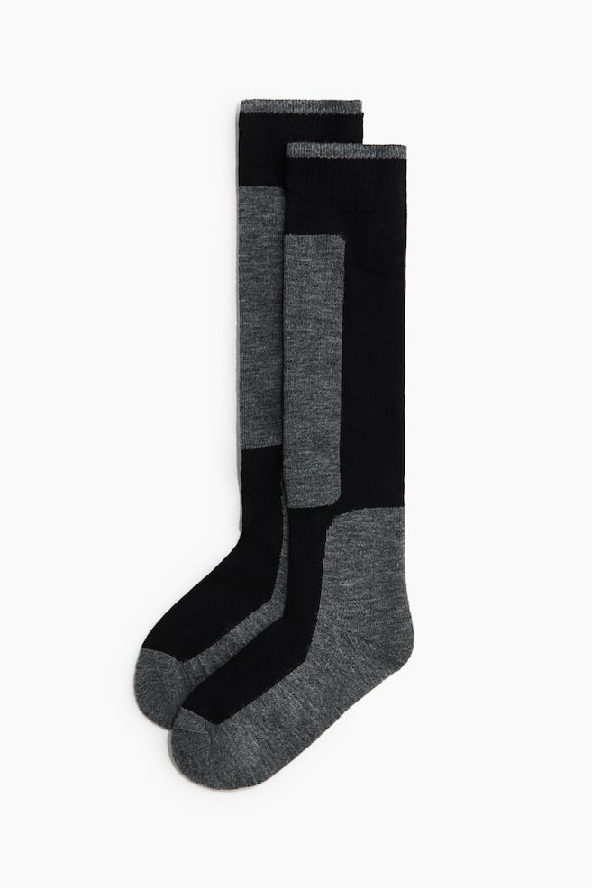 A pair of knee-high socks is laid flat, showcasing a colour-blocked design in black and heather grey. The socks feature ribbed cuffs, a vertical grey panel along the front of the shin, and contrasting grey heels and toes. - 1