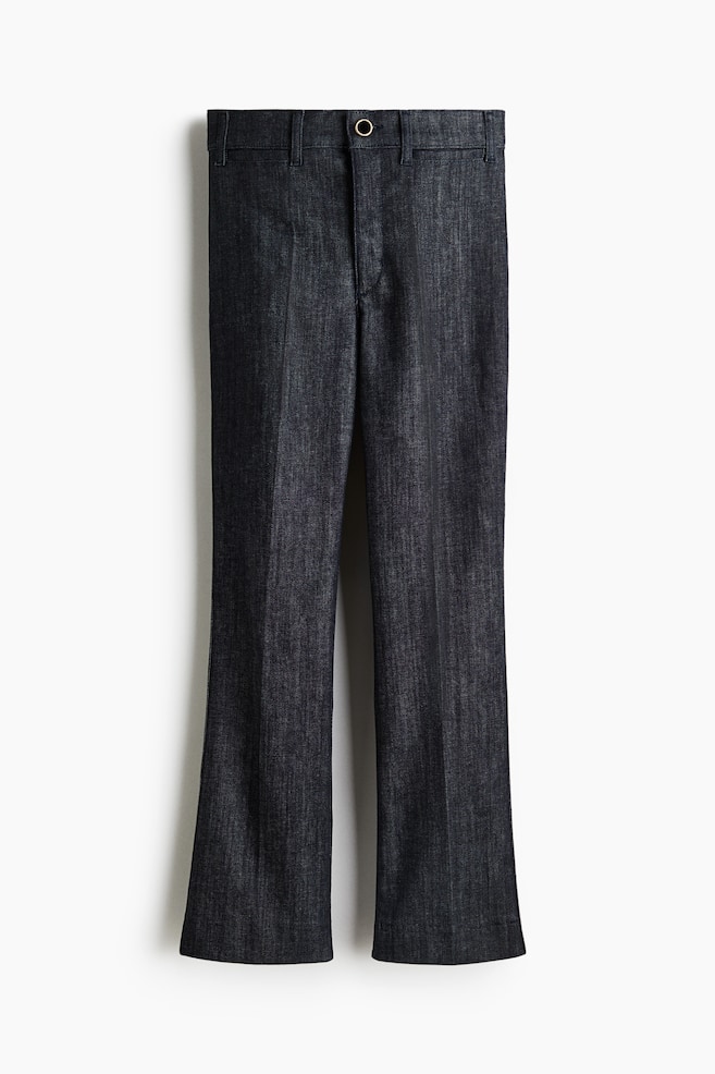 Dark wash denim pants are shown from the front, featuring a single silver-toned button and zipper fly closure at the waist. The pants are designed with a bootcut flare and a subtle vertical crease running down the front of each leg. - 1