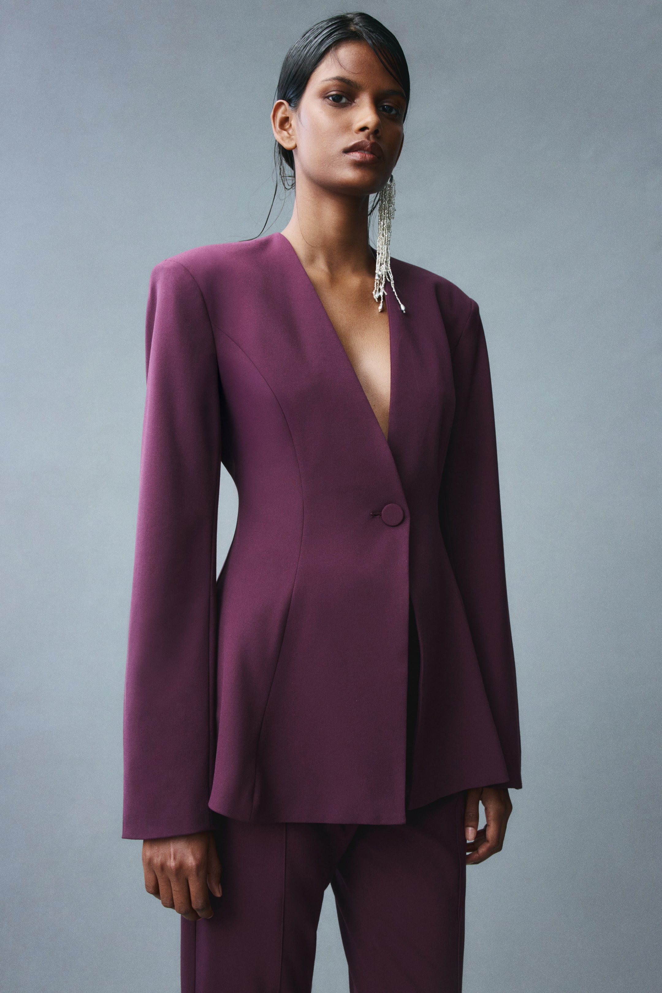 View larger image: The model is wearing a dark purple single-breasted blazer featuring a lapelless deep V-neckline, structured shoulder pads, and a single covered-button closure. The design includes princess seams that contour the waist before flaring out into a peplum hem, paired with matching trousers.