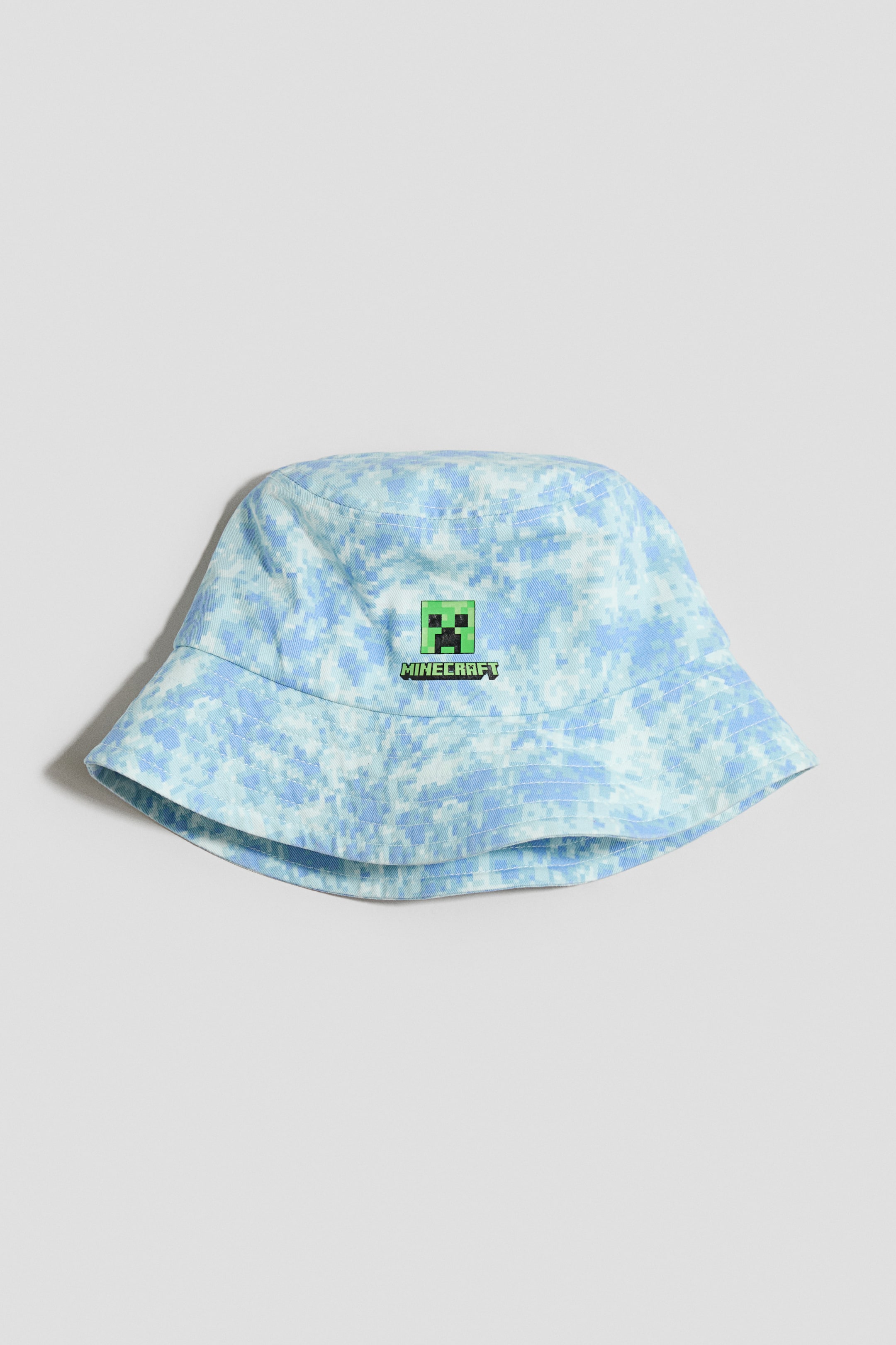 Näytä suurempi kuva: Ämpärihattu, jossa on vaaleansininen ja valkoinen pikselöity naamiointikuvio. Hatun etuosassa on vihreä pikselöity Minecraft Creeper -kasvologo, jonka alla on teksti "MINECRAFT".