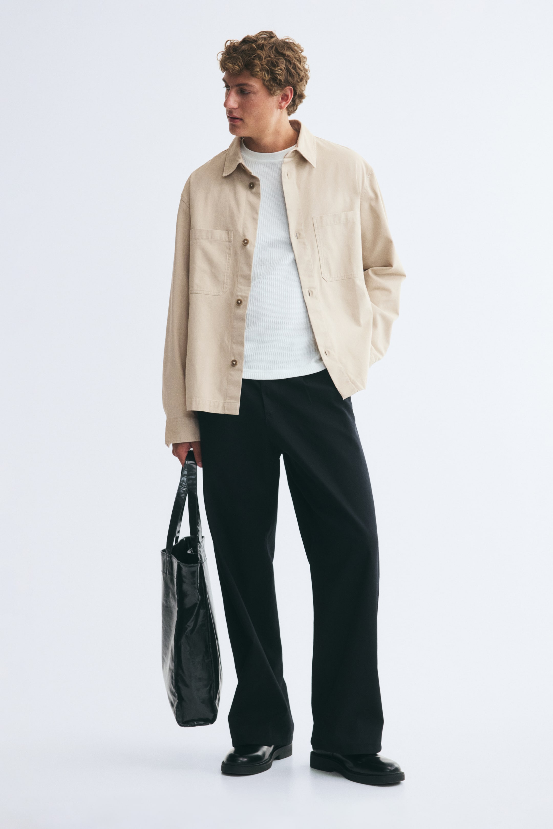 View larger image: A beige cotton twill overshirt with a collar, open chest pockets, and long sleeves with button cuffs is worn open by the model over a white T-shirt. The overshirt is styled with black trousers, and the model is holding a black tote bag.