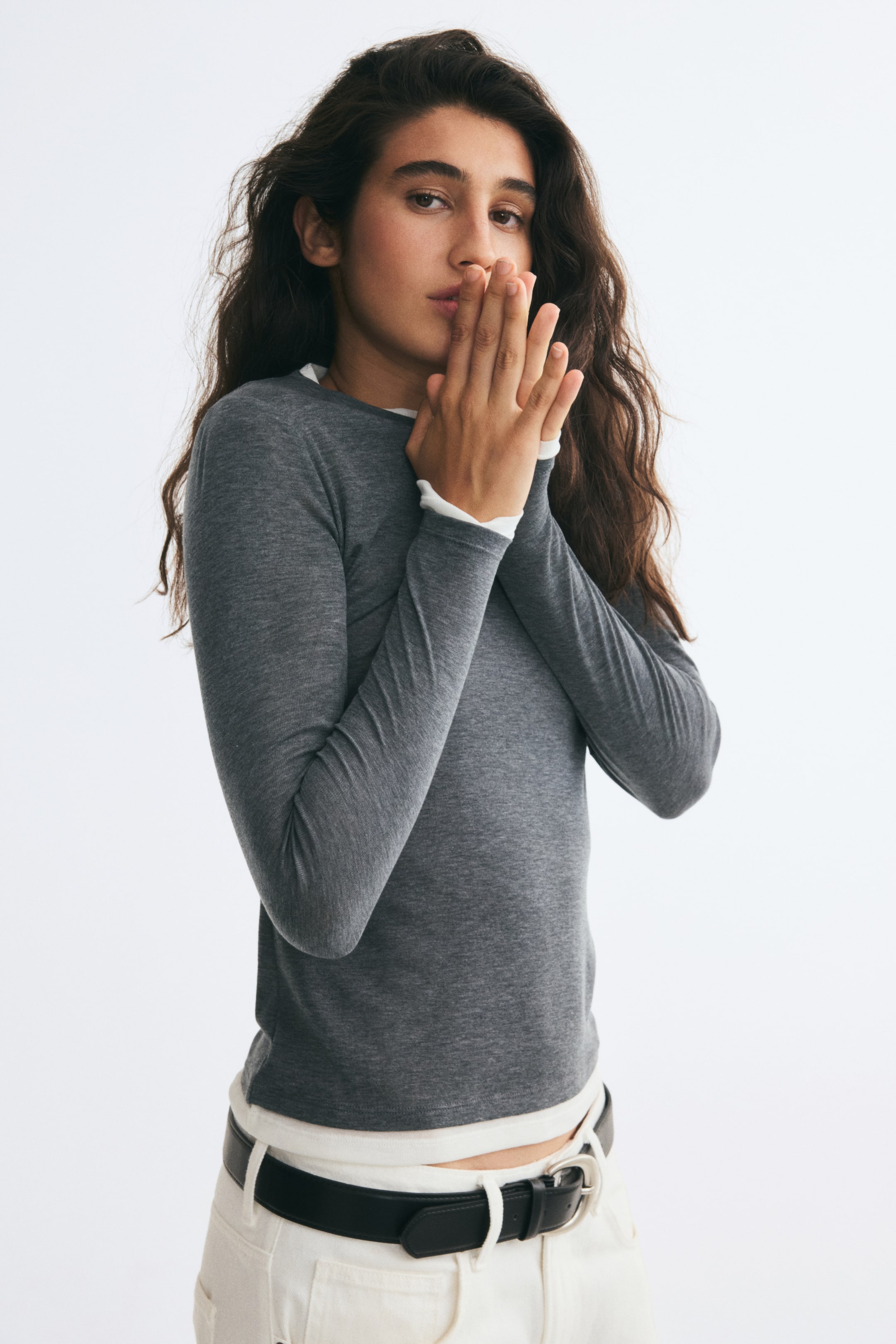 Ampliar la imagen: La modelo lleva una prenda de punto fino gris de manga larga, con ribetes blancos en contraste en el escote, los puños y el bajo. La modelo está de pie con las manos entrelazadas cerca del rostro, combinando la prenda con pantalones blancos y un cinturón negro.