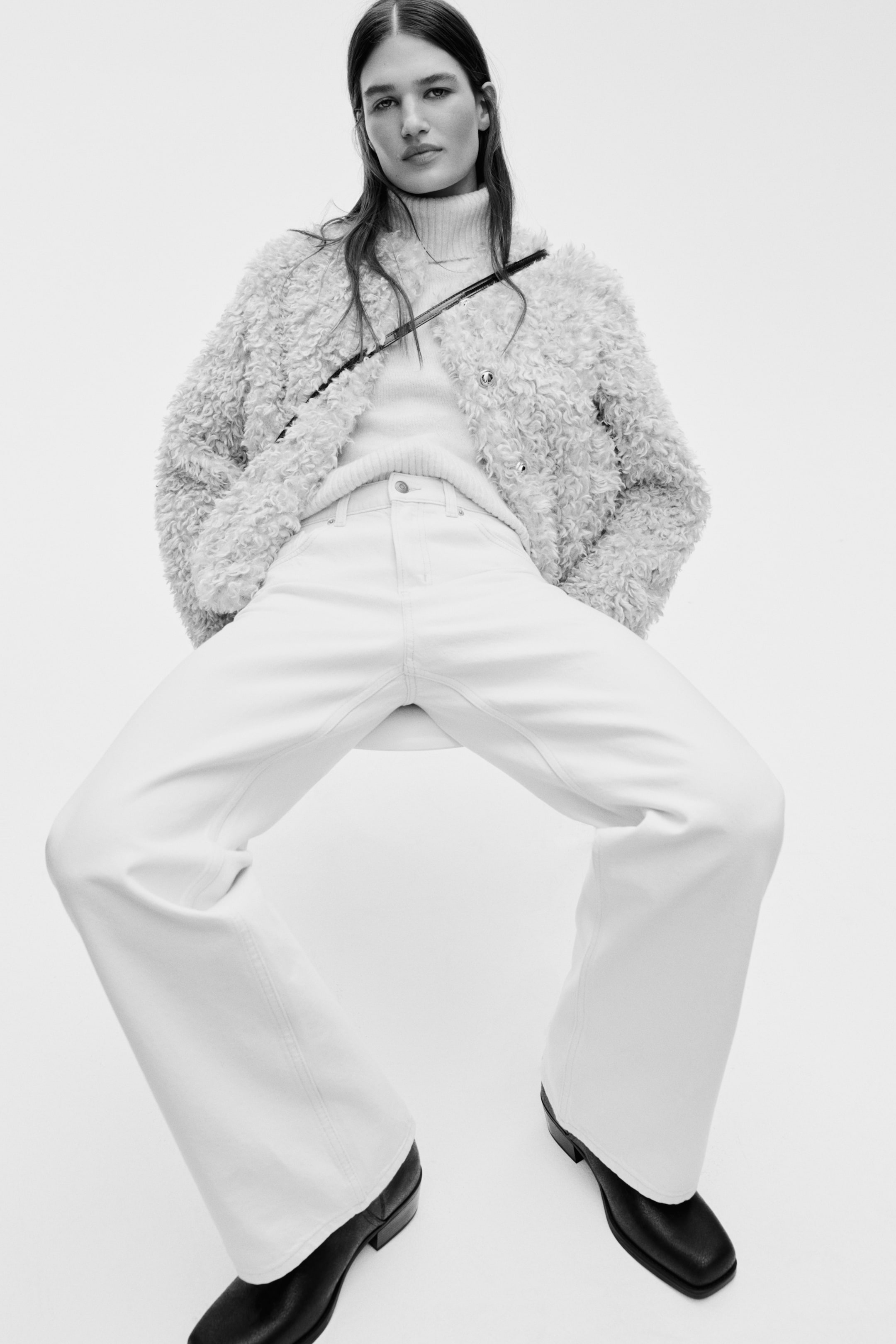 View larger image: An off-white, shaggy faux-shearling jacket layered over a ribbed turtleneck sweater, paired with matching wide-leg pants featuring prominent front seams that run down the legs. Black square-toed boots peek out from beneath the pants, all presented from a low, frontal angle.