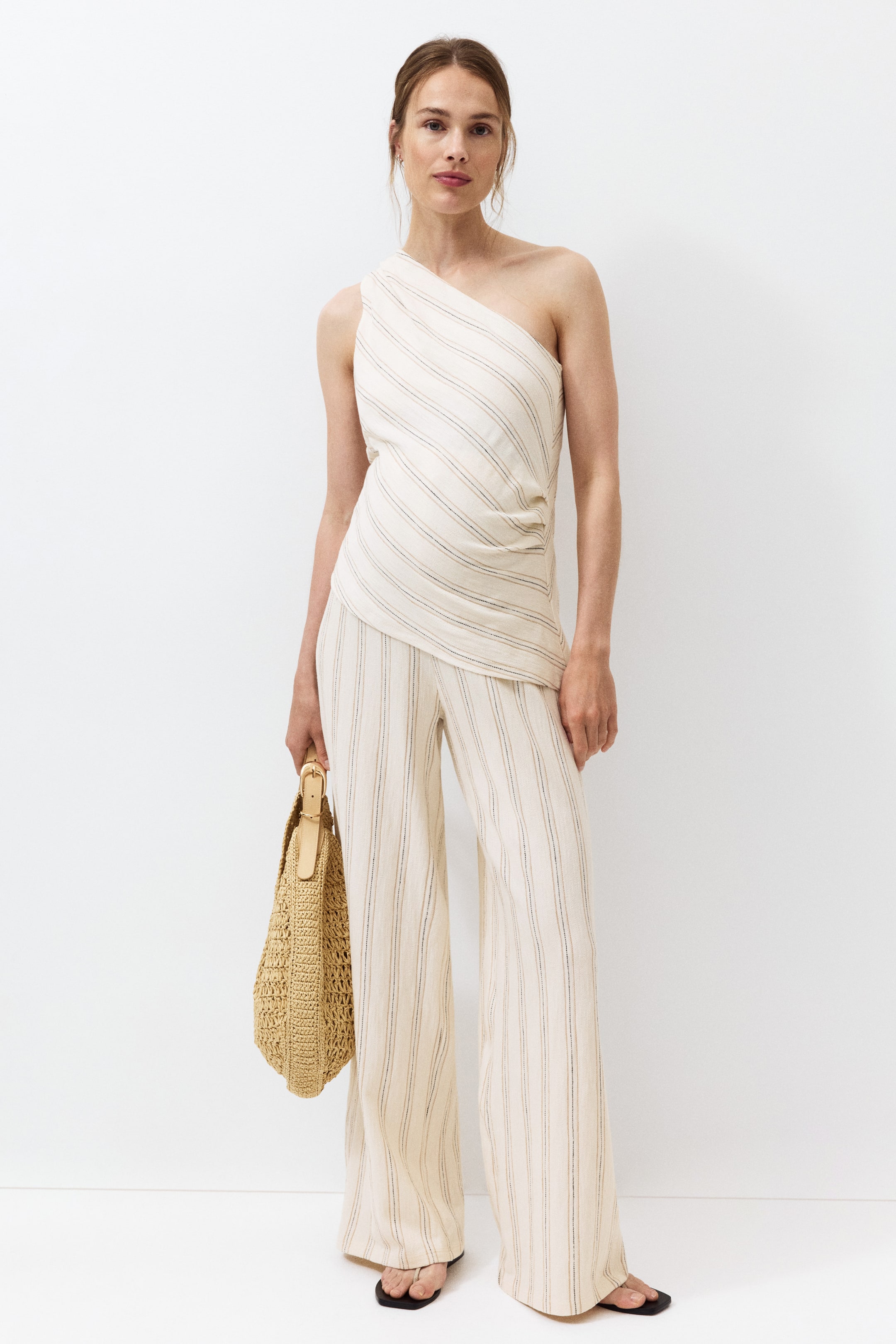 Agrandir l'image: Un haut asymétrique blanc cassé avec des rayures fines diagonales et un drapé latéral est présenté de face, associé à un pantalon large assorti avec des rayures fines verticales. Un sac à bandoulière en paille tressée est tenu, et des sandales noires à lanières en forme de tongs avec un bout carré sont portées.