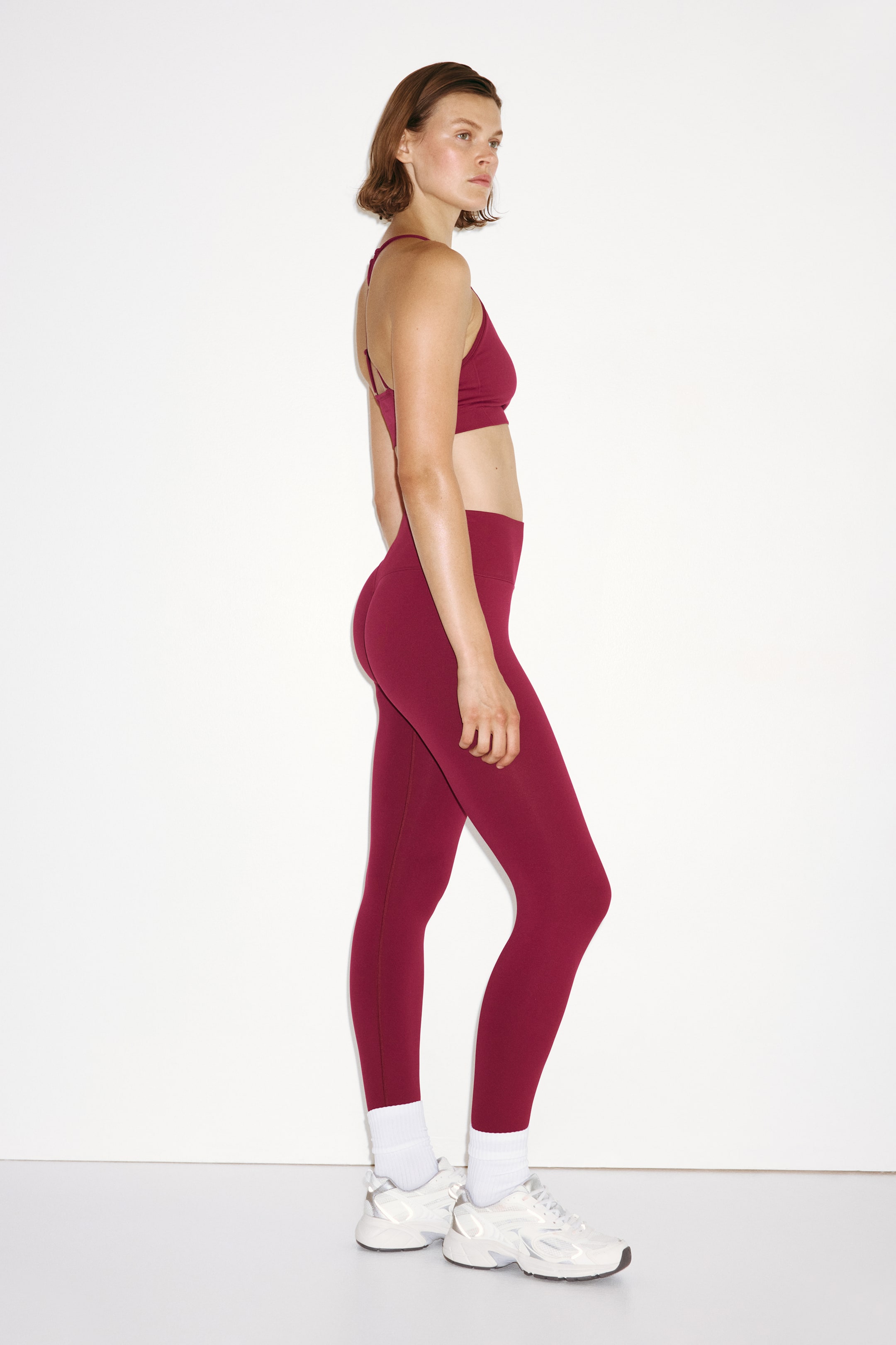 Grotere afbeelding bekijken: Naadloze sportlegging met DryMove? in dieprood wordt gedragen door het model. De hoog uitgesneden, aansluitende, lange legging wordt gecombineerd met een bijpassende sportbeha, witte crew sokken en witte sportsneakers.