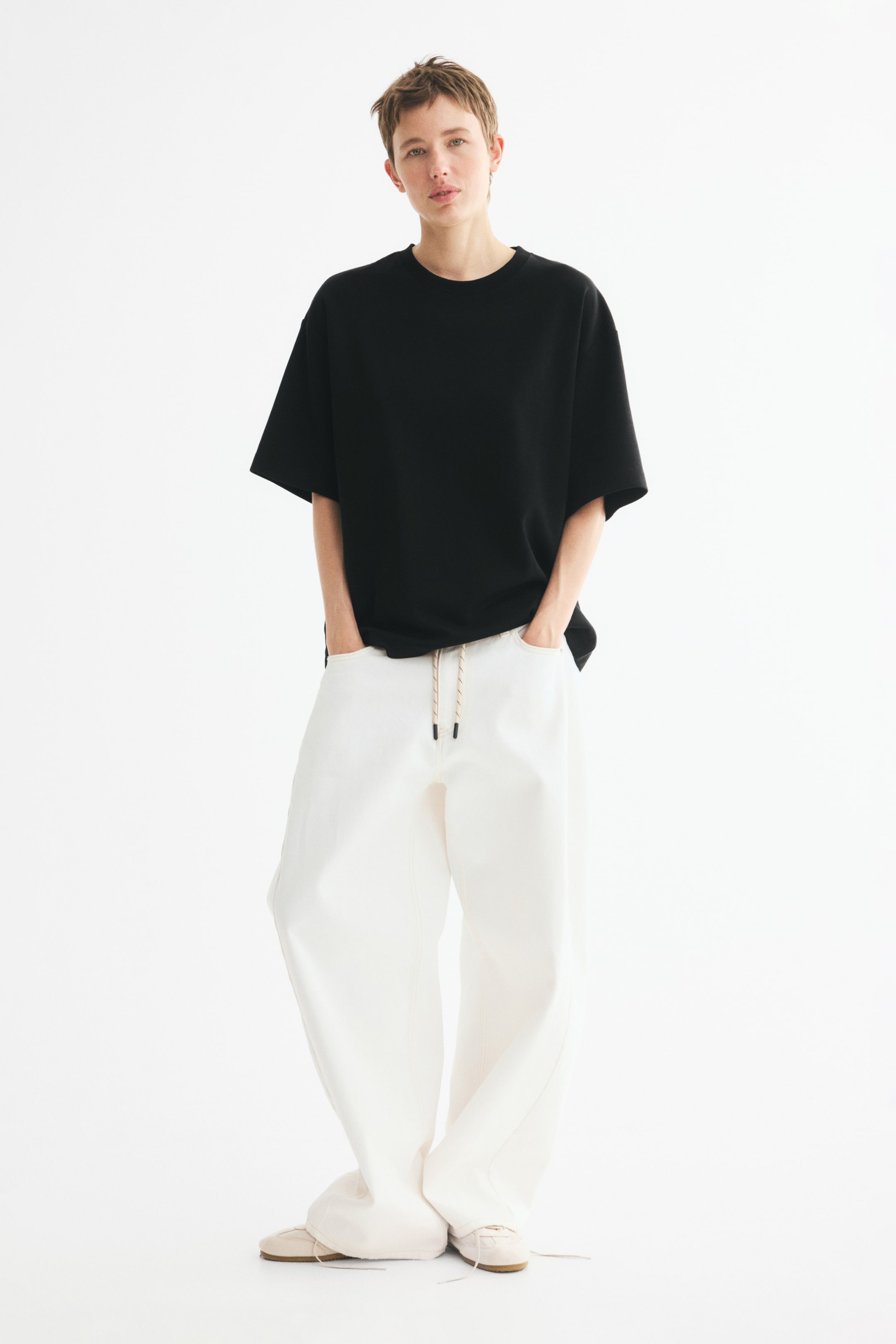 View larger image: The model is wearing a black loose-fit T-shirt in heavy interlock jersey, featuring a round rib-trimmed neckline and dropped shoulders. The model is standing with hands placed in the pockets of wide-leg white trousers styled with the tee.