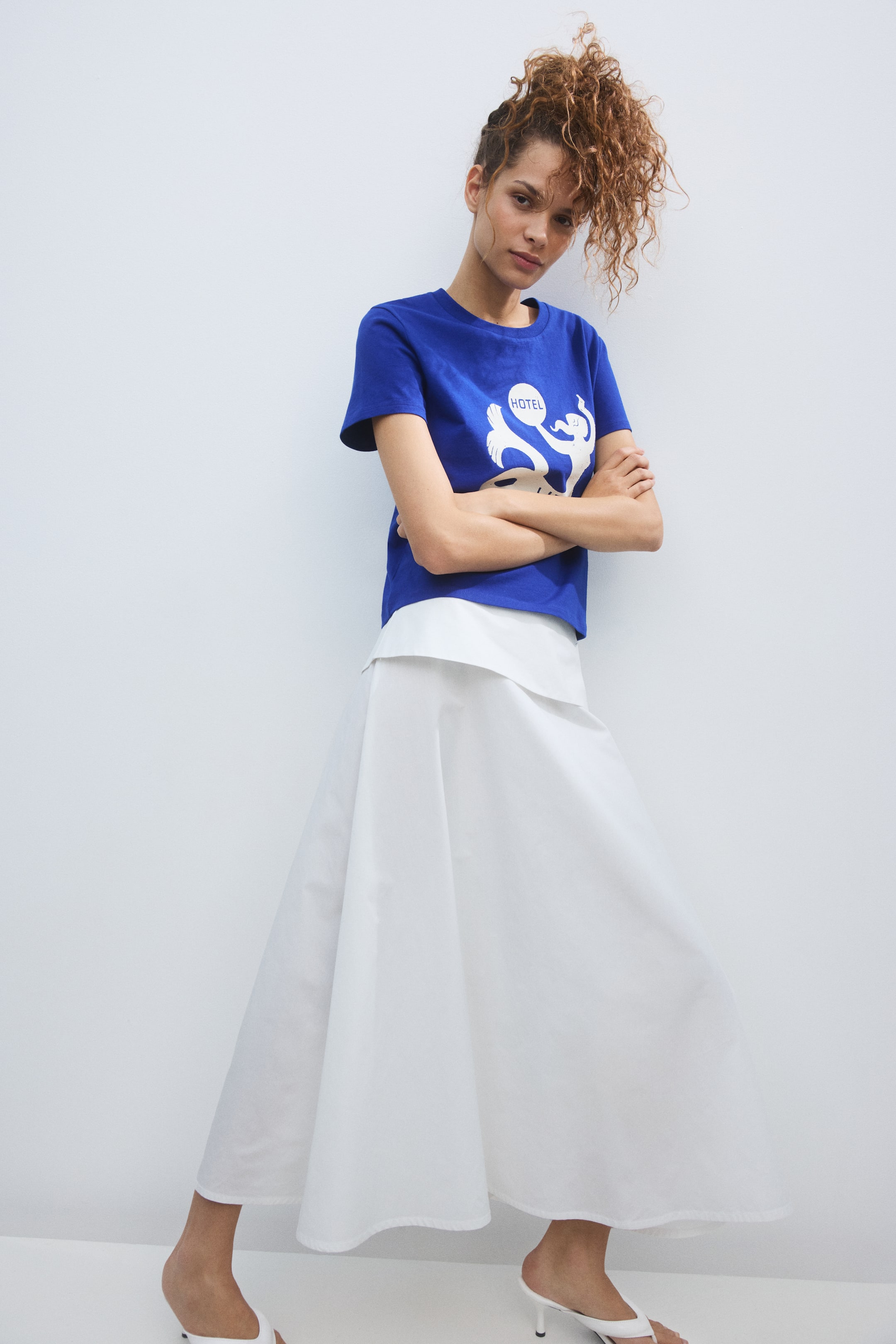 Zobrazit větší obrázek: Královsky modré tričko s kulatým výstřihem a krátkými rukávy s bílým grafickým potiskem je nošeno přes bílý zkrácený top, který je kombinován s bílou midi sukní.