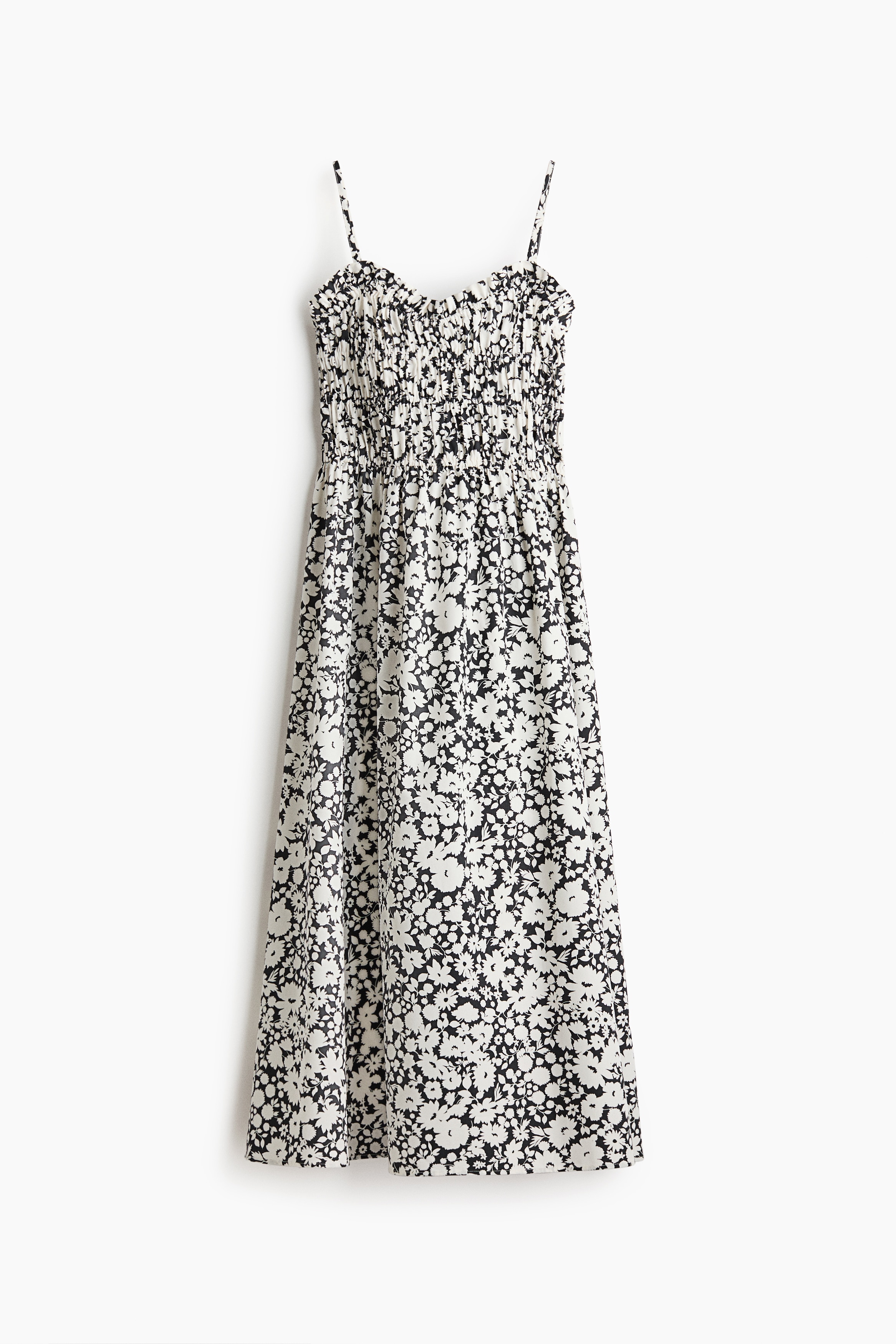 A calf-length dress featuring a black and white floral print, thin shoulder straps, a ruched bodice, and a flared skirt is being worn by the model. The model is also holding a small black handbag and wearing black flat shoes.