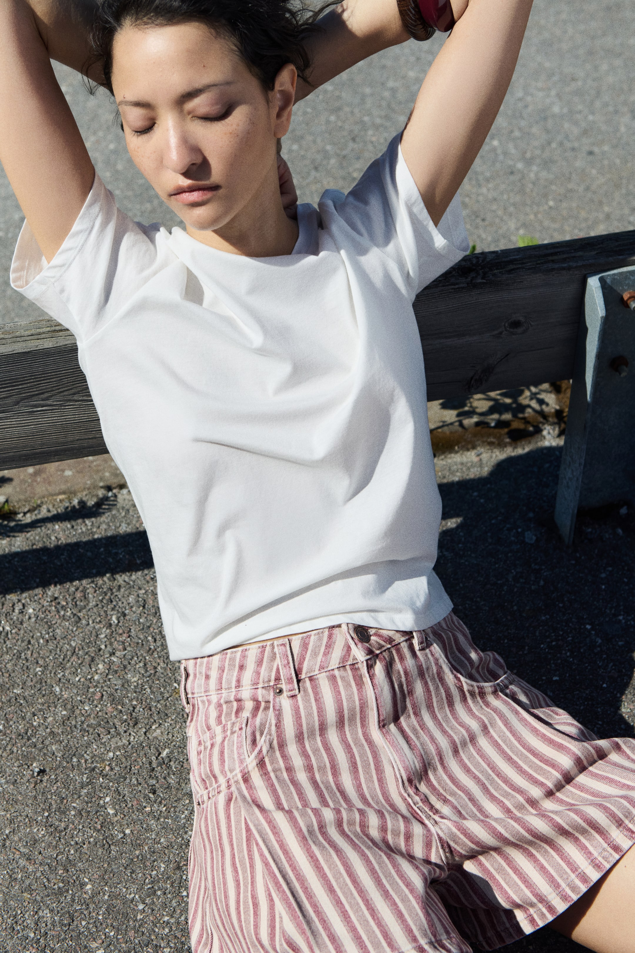 Visualizza l'immagine ingrandita: Una t-shirt bianca a girocollo presenta maniche corte e una vestibilità rilassata, indossata infilata in pantaloncini a vita alta. I pantaloncini mostrano strisce verticali color marrone e crema, insieme a tasche frontali con rivetti e passanti per cintura visibili.