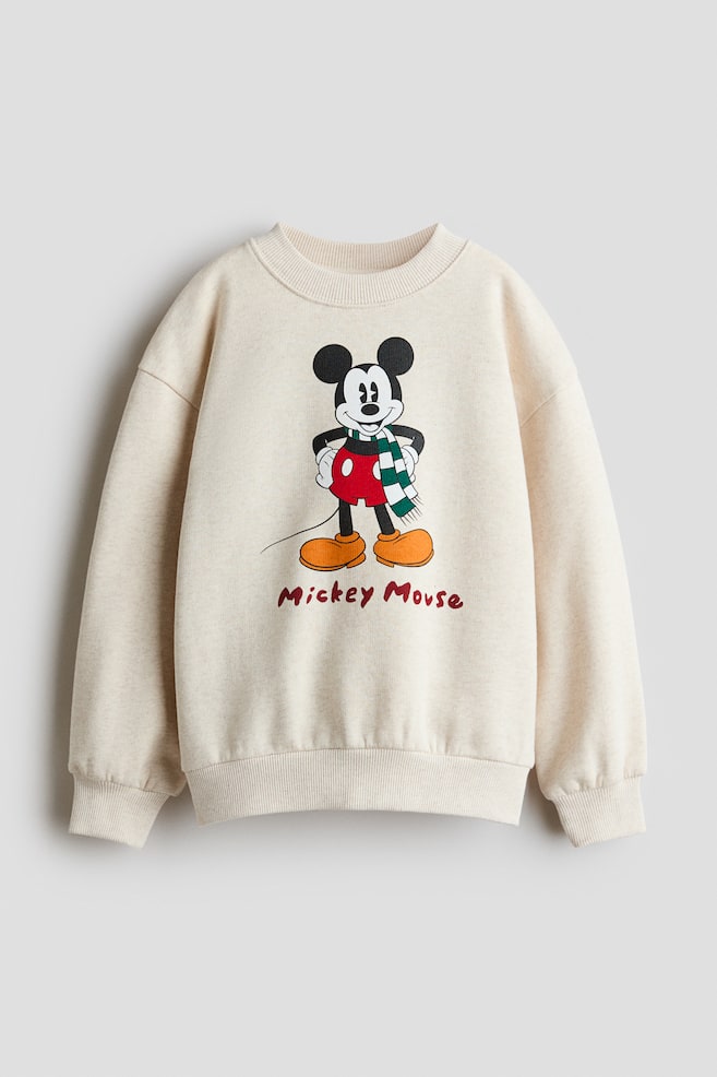 A cream-coloured fleece sweatshirt shown from the front features a printed graphic of Mickey Mouse wearing a green-and-white striped scarf on the chest, with the name "Mickey Mouse" printed below. The garment has a rib-trimmed round neckline, dropped shoulders, and ribbed cuffs and hem. - 1