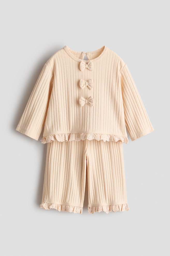 A light beige ribbed two-piece set is presenting, consisting of a long-sleeved top and matching pants. The top features three decorative bows vertically aligned on its FRONT, and both the top's hem and the pants' leg openings are adorned with a ruffled lace trim. - 1