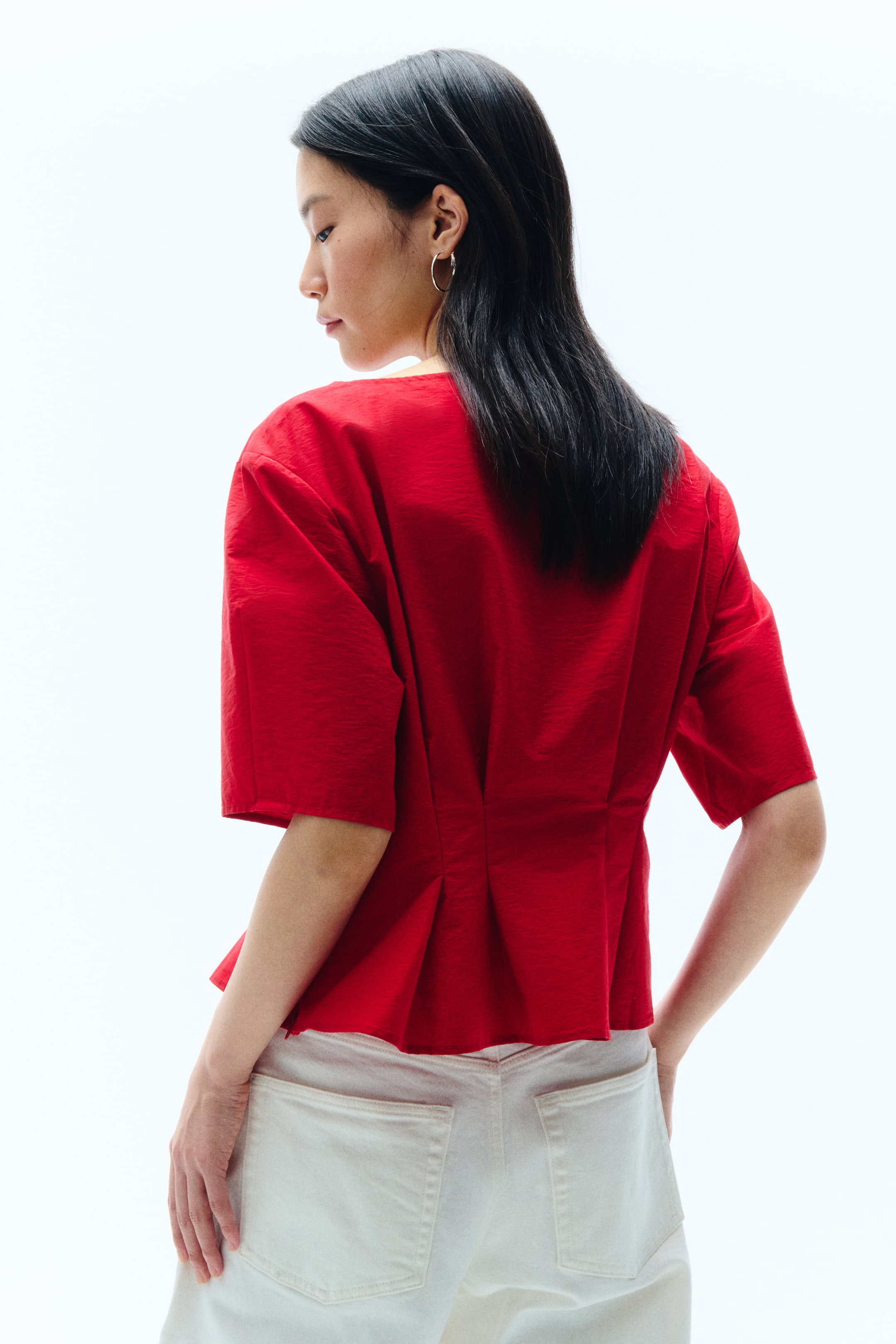 View larger image: A red top features a slightly textured fabric, short wide sleeves, and a gathered peplum hem at the back that creates a flared silhouette. It is styled with white pants, showing its cropped length from a rear-side angle.