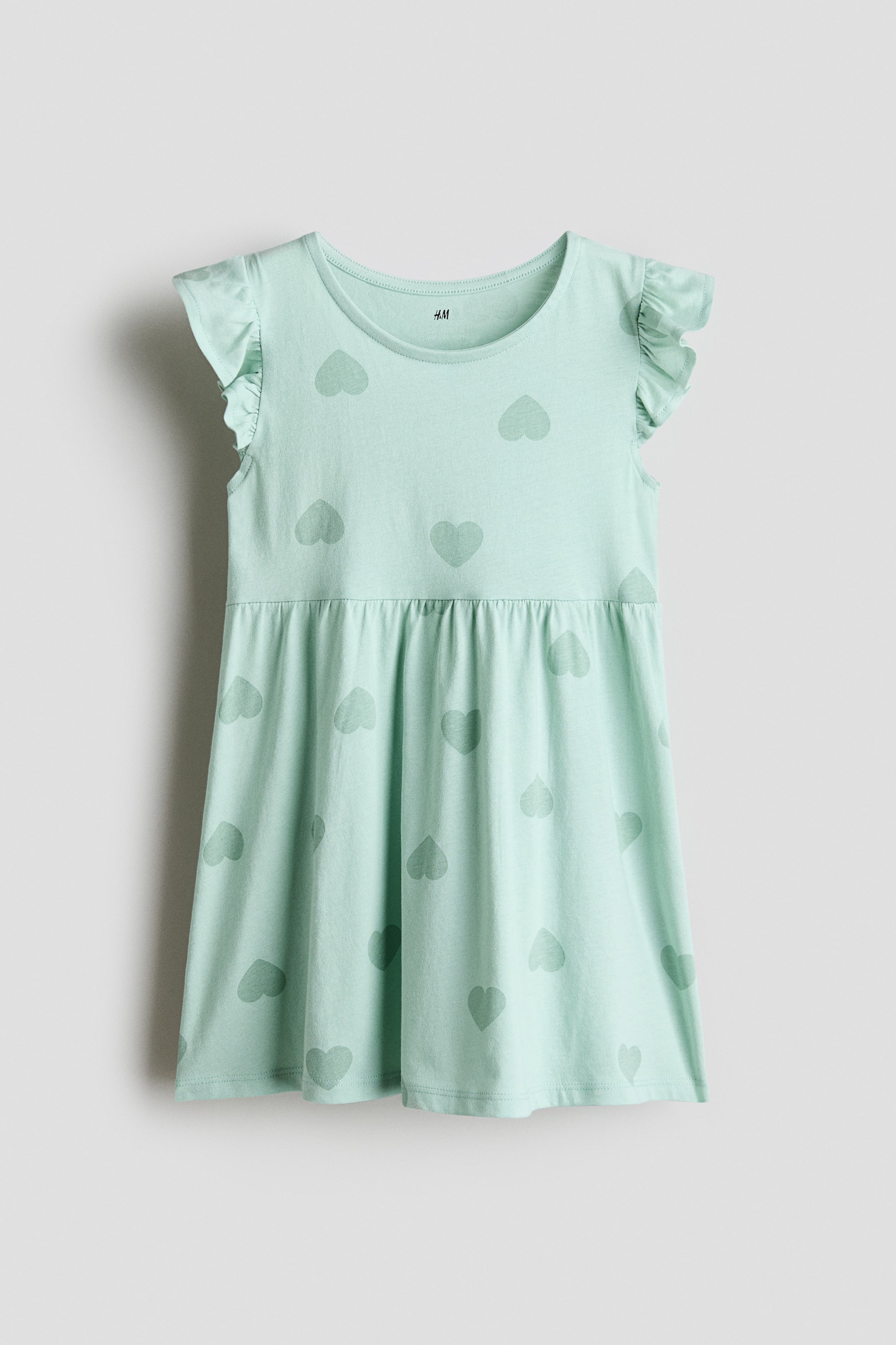 View larger image: A light mint-green dress in soft cotton-jersey is presented from the FRONT. It features a round neckline, flutter sleeves and a gathered seam at the waist, with a subtle tonal heart pattern visible throughout.