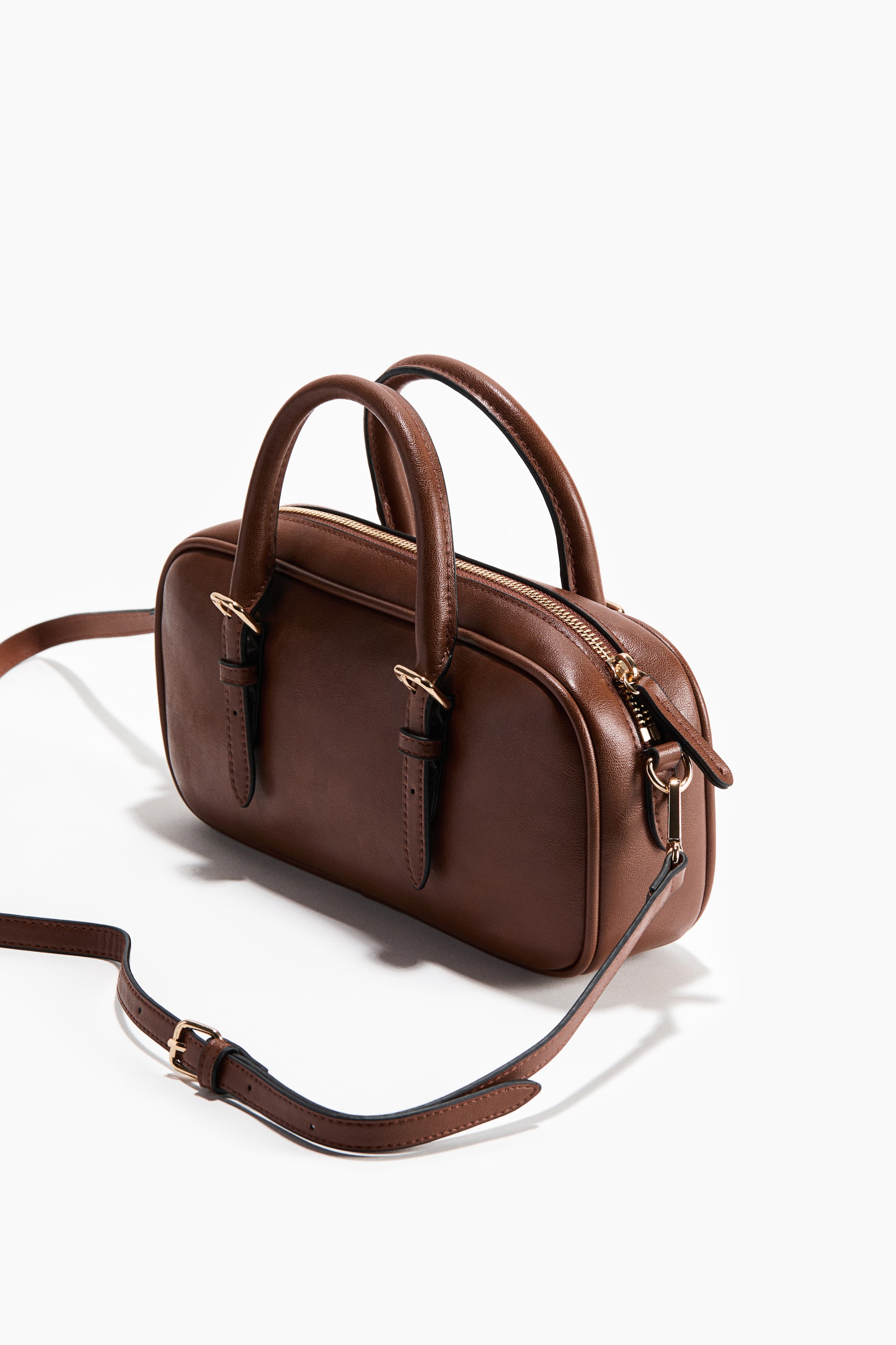 查看大圖: 一款棕色細長手提包,配有兩個圓弧形頂端提把及帶有金色拉片的頂部拉鍊。側邊延伸出可調式肩帶,肩帶與提把的連接處均飾以金色五金扣。