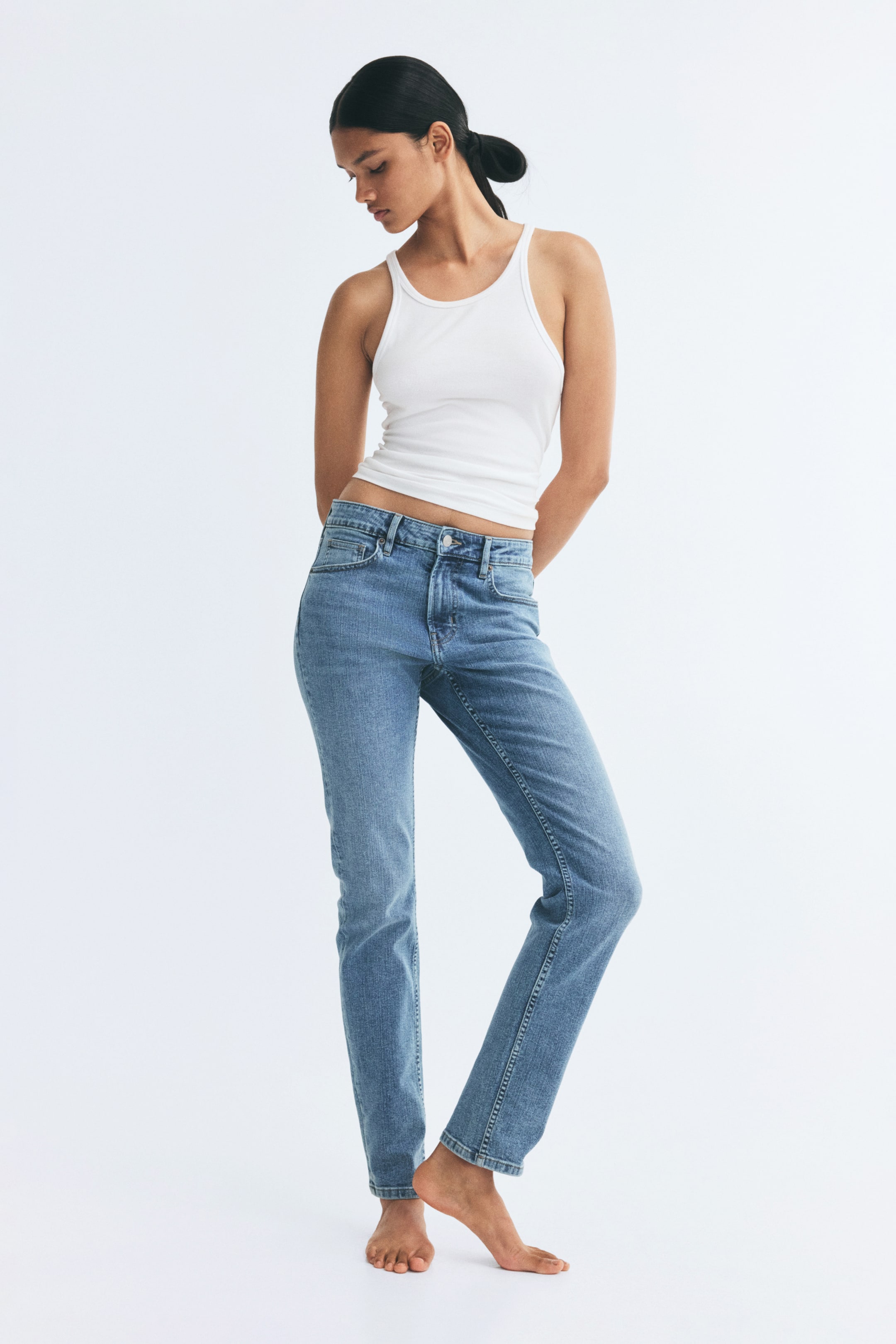 View larger image: The model is wearing five-pocket jeans in a medium blue wash, paired with a white ribbed tank top. The jeans feature a regular-rise waist with a button closure and a slim fit through the legs.