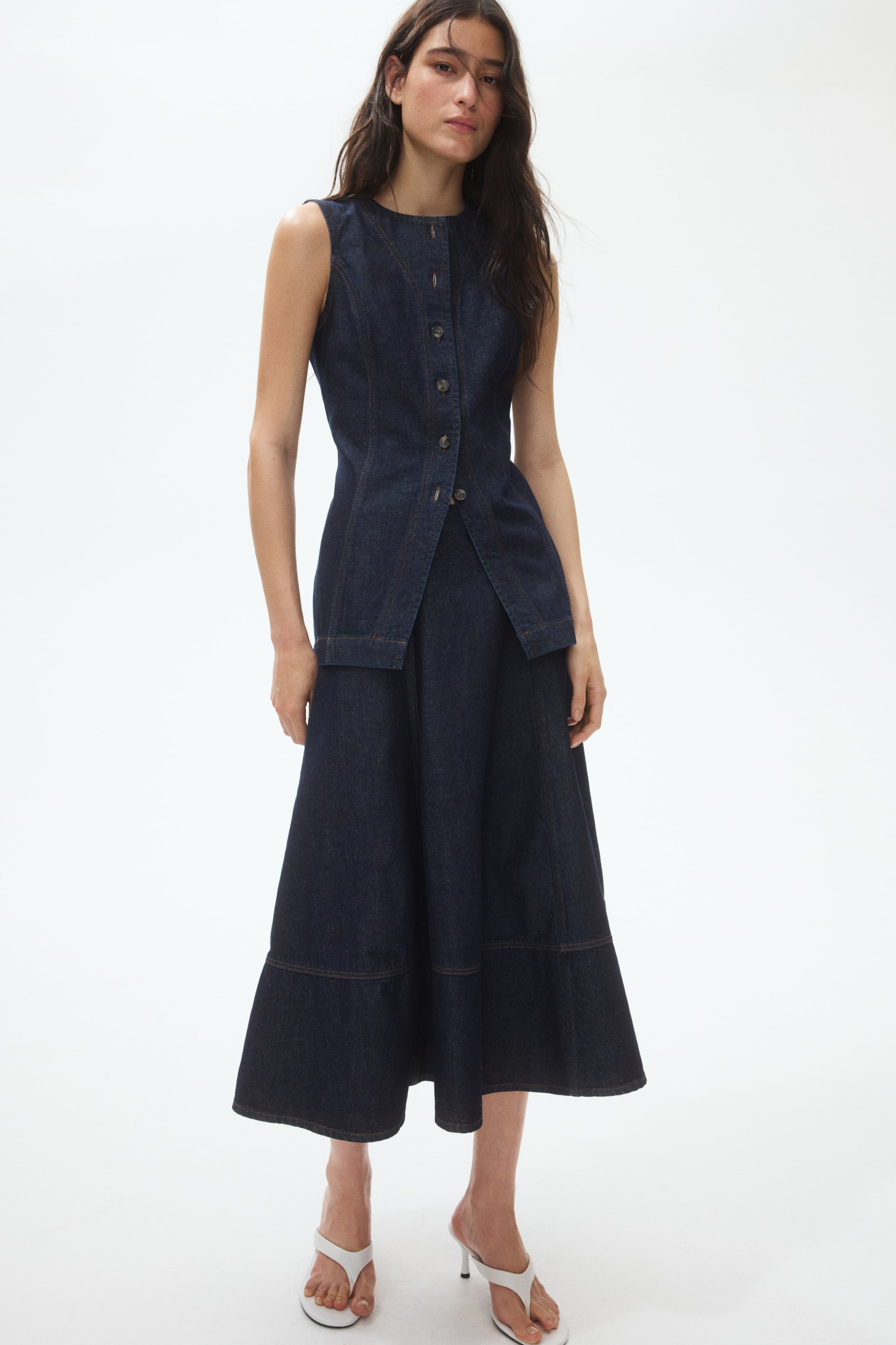 View larger image: A dark wash denim sleeveless top and matching midi skirt are showcased directly. The top features a round neck, button-front closure, and a structured, split hem, while the skirt displays a flared silhouette with a tiered panel near the hem.