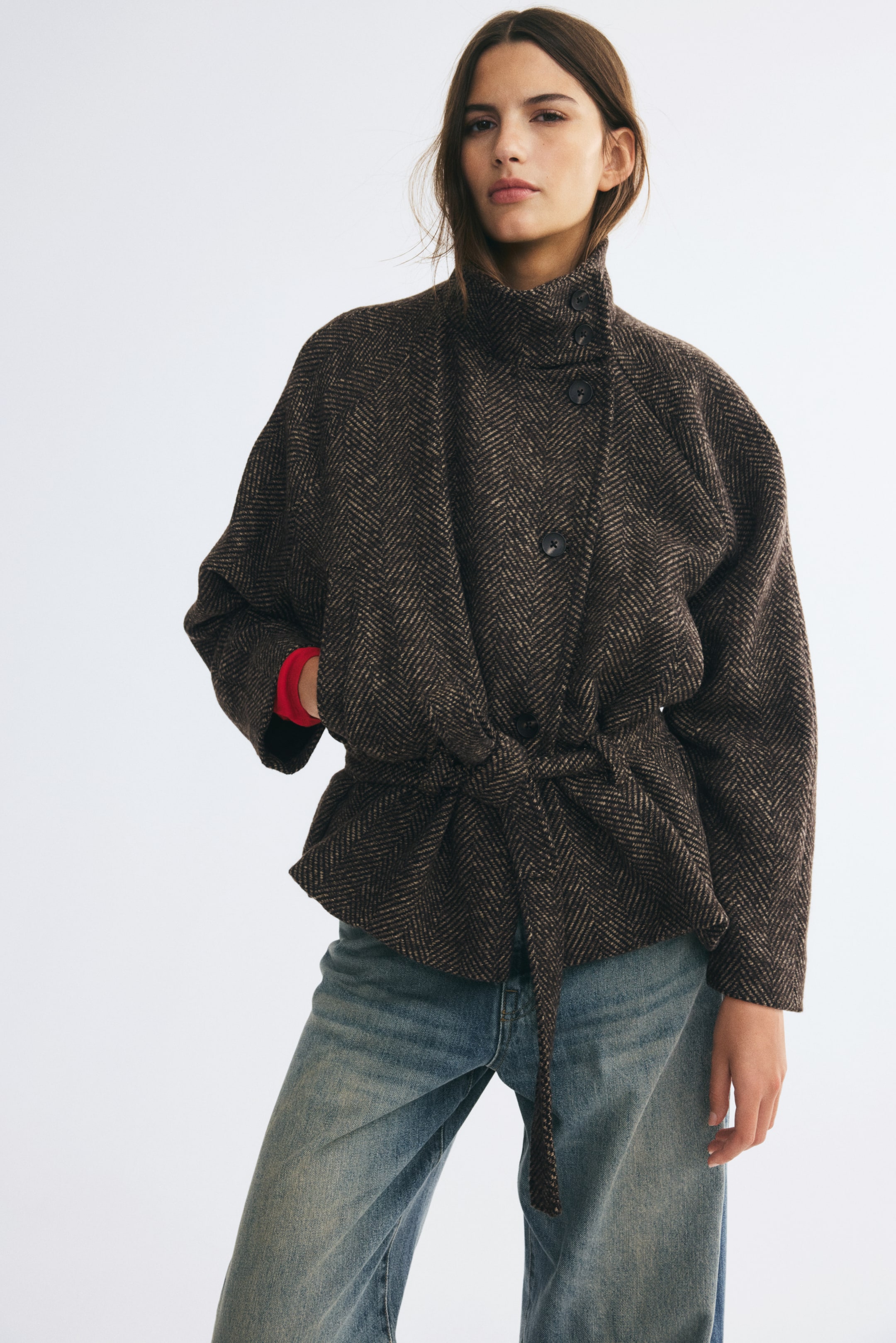 Ver imagen más grande: La modelo lleva una chaqueta de tejido texturizado marrón con estampado espiga, con cuello mao abotonado, cierre delantero asimétrico con botones y un ancho cinturón de lazo en la cintura. La modelo apoya una mano en un bolsillo de vivo delantero, dejando ver un puño rojo que asoma de la larga manga raglán, y combina la chaqueta con unos vaqueros azules.