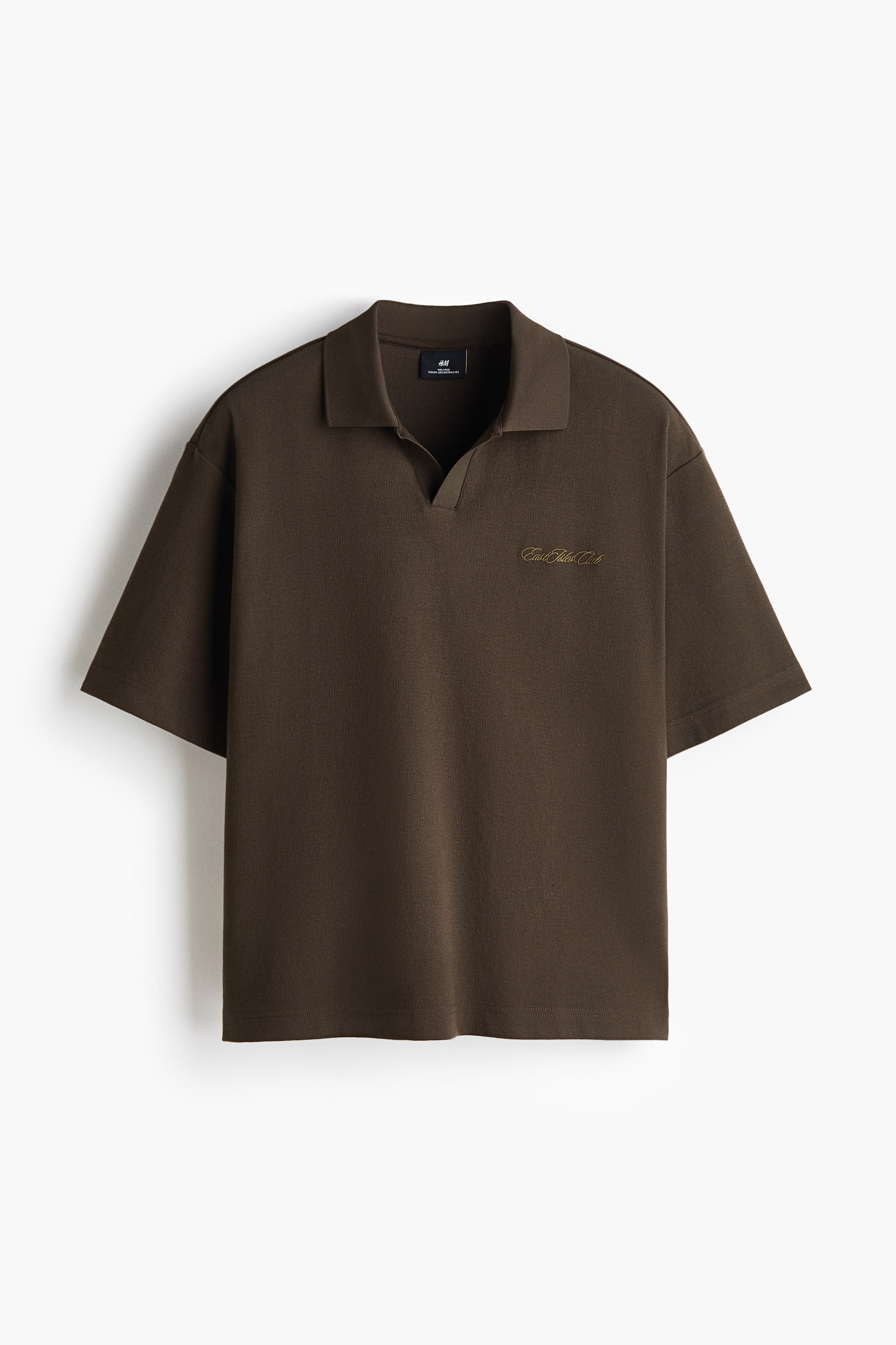 View larger image: A dark brown polo shirt is shown from the front, featuring a relaxed fit with dropped shoulders and wide short sleeves. It has a classic flat collar with a buttonless, open V-neck placket and a small gold-toned embroidered script on the left chest.