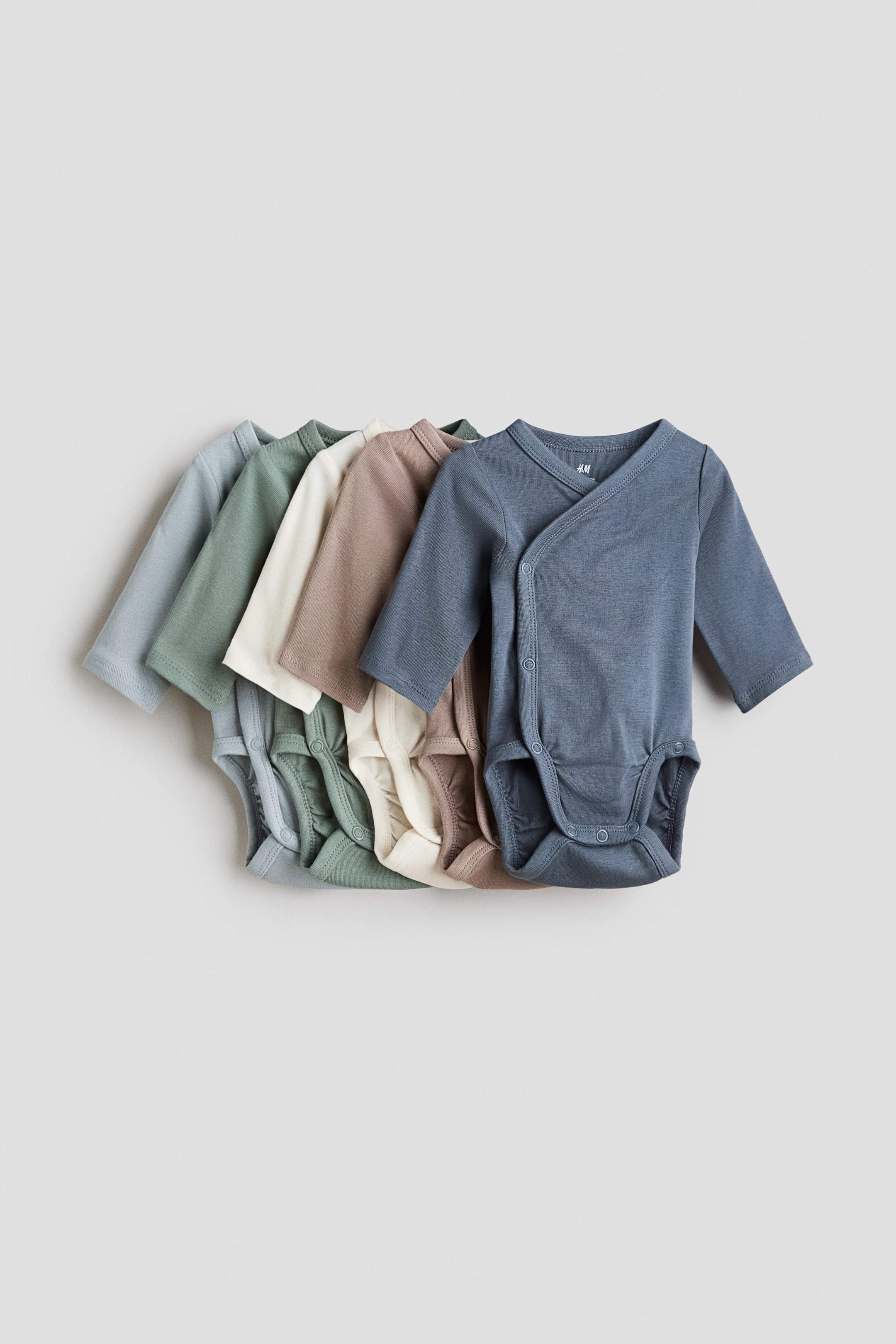 View larger image: Five long-sleeved bodysuits are arranged in a layered display, showcasing muted shades of light blue, sage green, cream, light brown, and blue-grey. The frontmost bodysuit, in blue-grey, features a smooth, jersey-like wrapover front with press-studs at the sides and bottom, and gathered seams are visible around the legs.