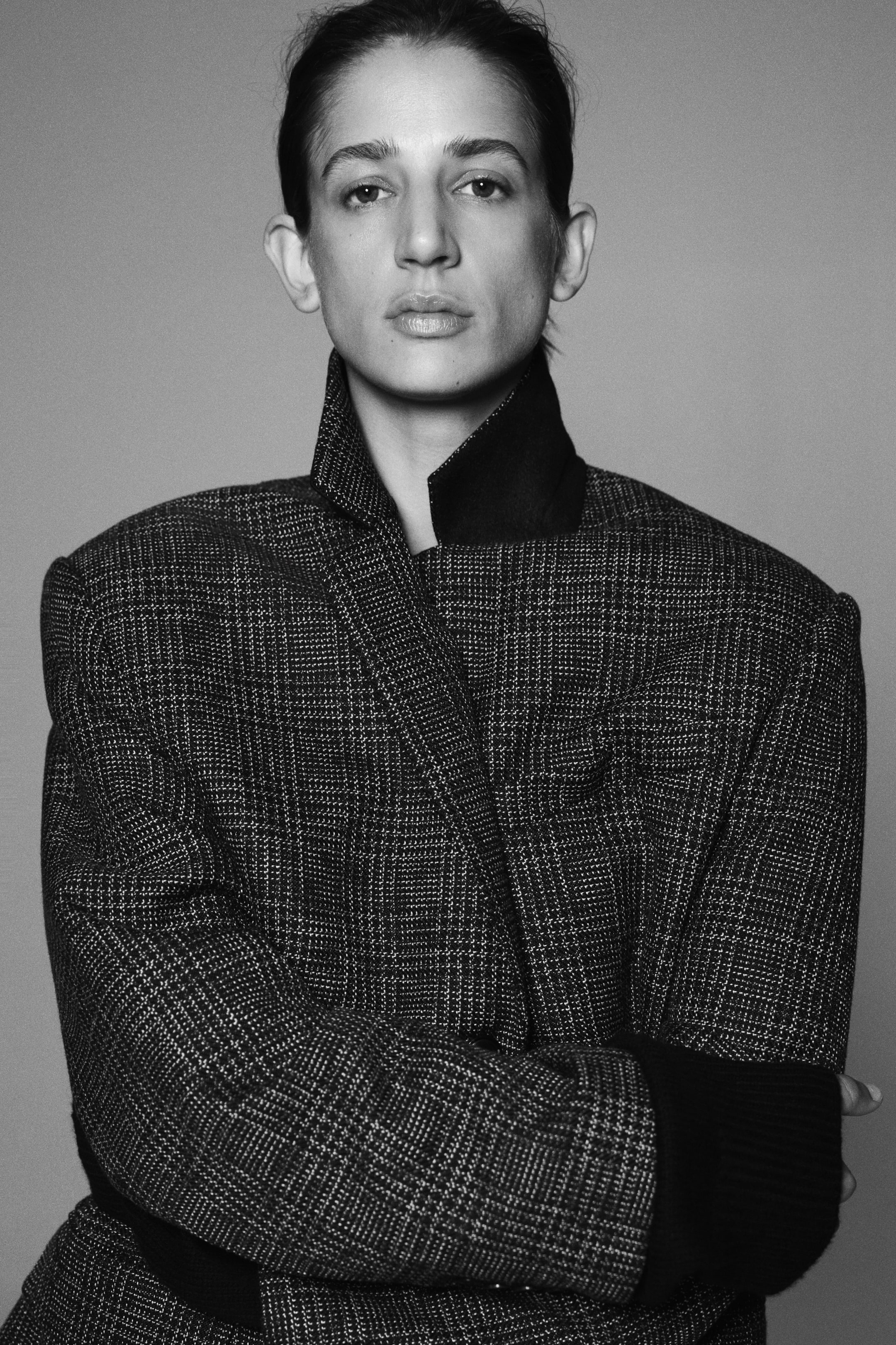Ampliar la imagen: La modelo lleva un blazer de un solo botón, con tejido texturizado y un patrón oscuro, mientras cruza los brazos al frente. El blazer presenta hombros estructurados y cuello levantado, y se combina con una prenda de manga larga con un puño acanalado oscuro visible.