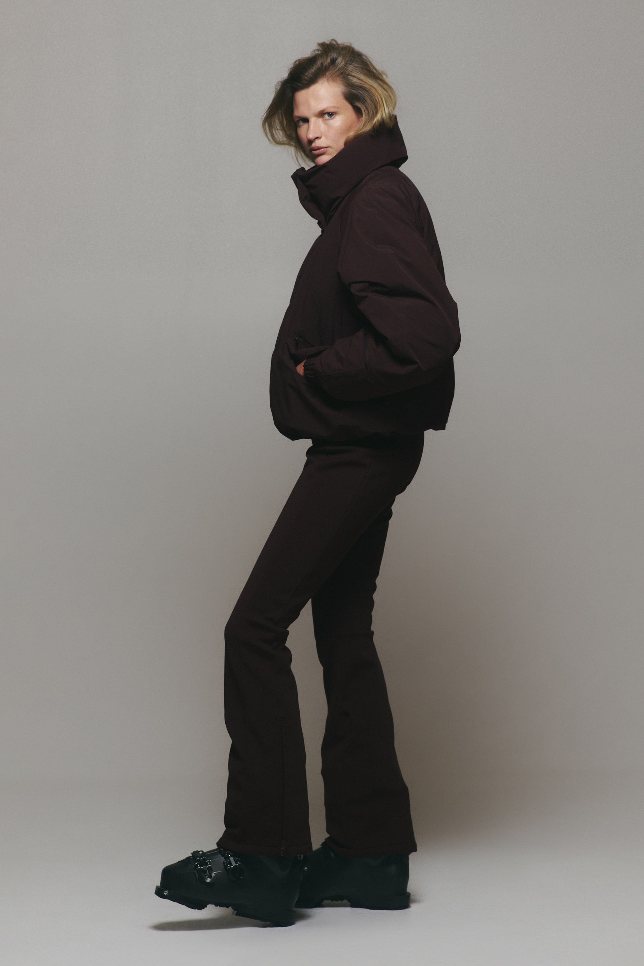 View larger image: A dark brown ski suit, consisting of a high-collar padded jacket and matching slim-fit trousers, is being worn by the model, who is standing in profile with hands tucked into the jacket pockets. The outfit is styled with black ski boots.