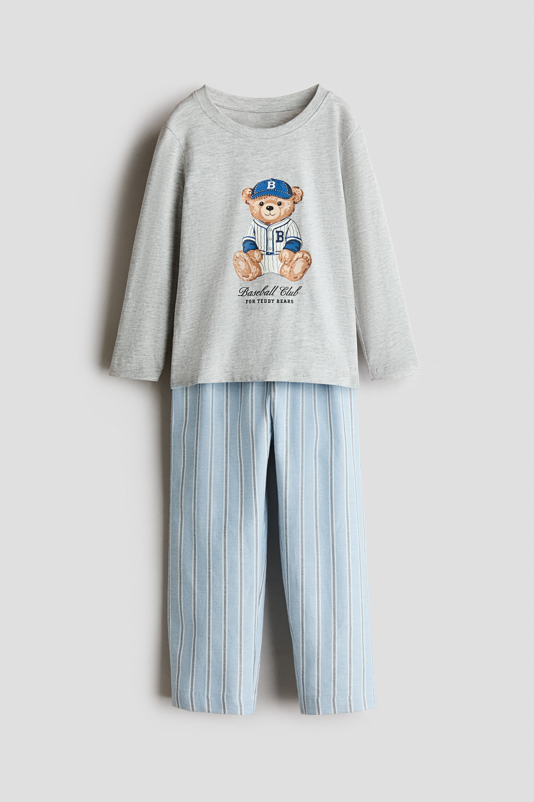 View larger image: A children's pajama set is on display, featuring a grey long-sleeved top and light blue striped pants. The top showcases a graphic print of a teddy bear wearing a blue baseball cap and a striped baseball jersey, with the text 'Baseball Club FOR TEDDY BEARS' below it, and the pants are designed with vertical grey and white stripes.