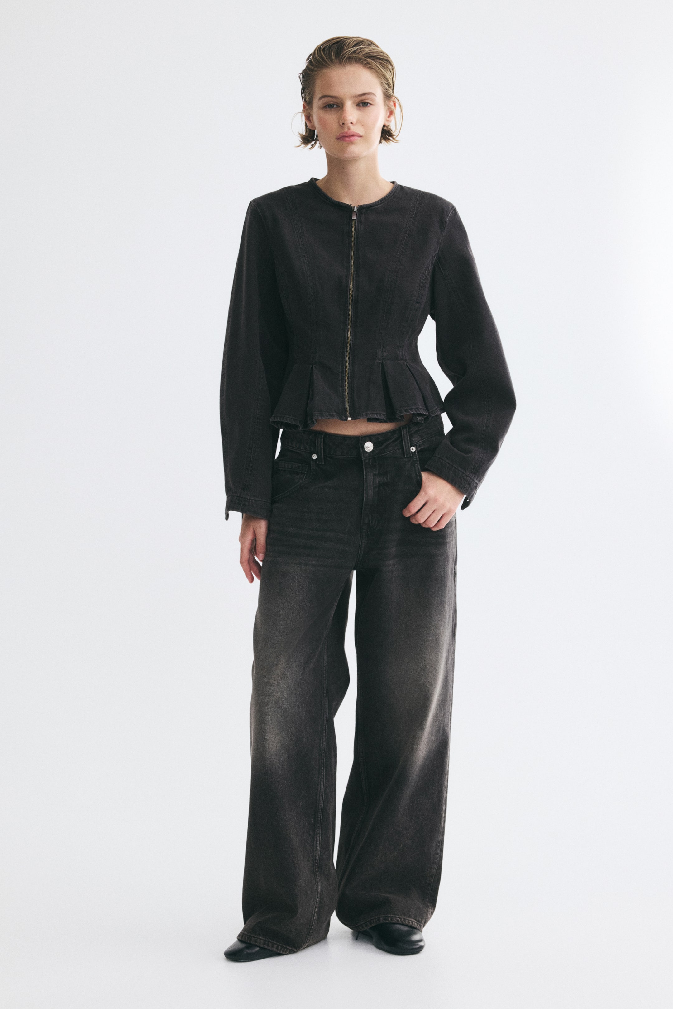 View larger image: The model is wearing faded black denim jeans with a low rise and a loose, wide-leg fit, standing with one hand in a front pocket. The jeans are styled with a matching black denim zip-up jacket featuring a peplum hem.