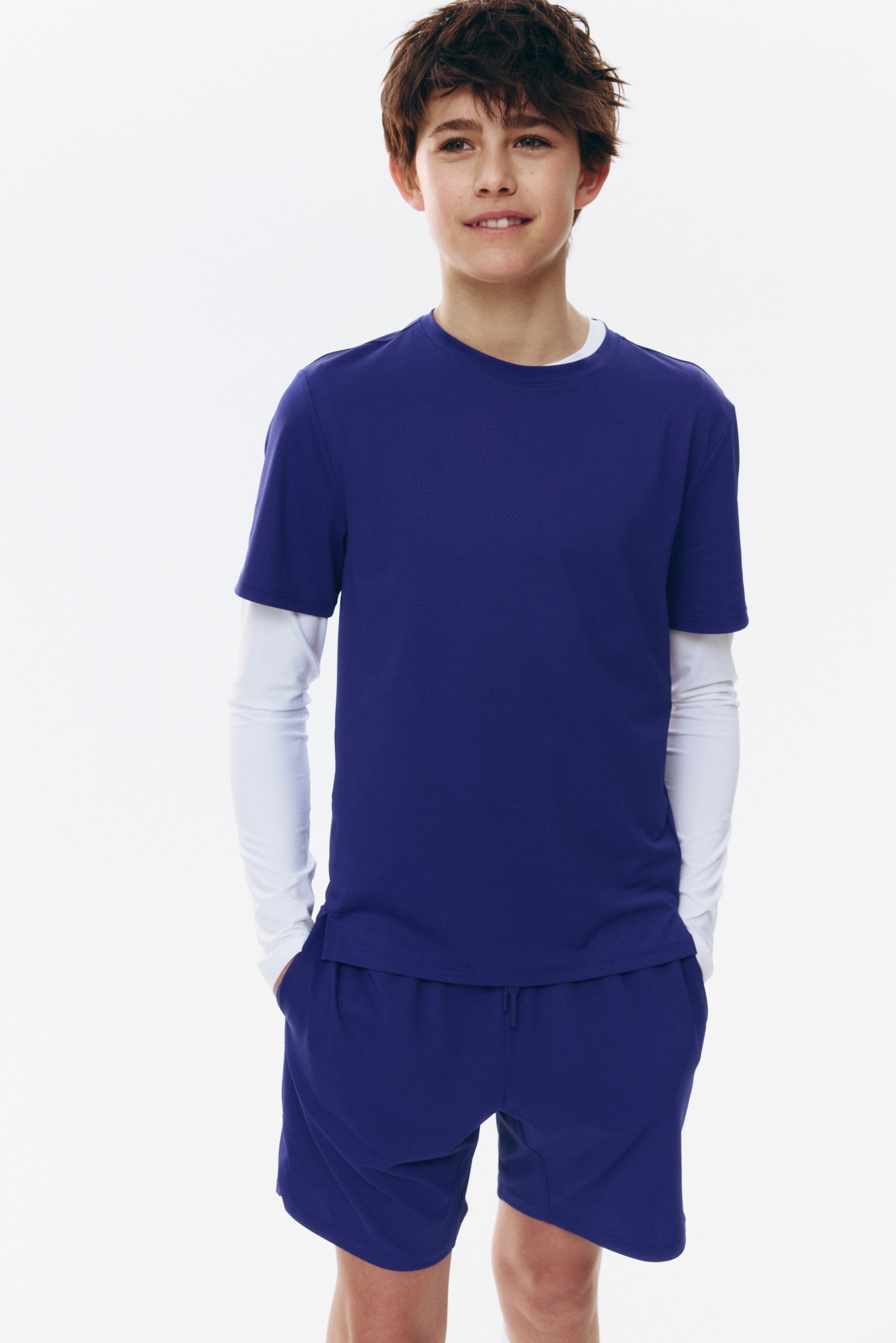Visa större bild: En pojkes sportiga outfit är vänd framåt, med en djupt lila, kortärmad, rundhalsad t-shirt med en texturerad yta, lagrad över en vit långärmad skjorta. Outfiten kompletteras med matchande djupt lila shorts, som visar ett synligt dragsnöre i midjan och sidofickor.