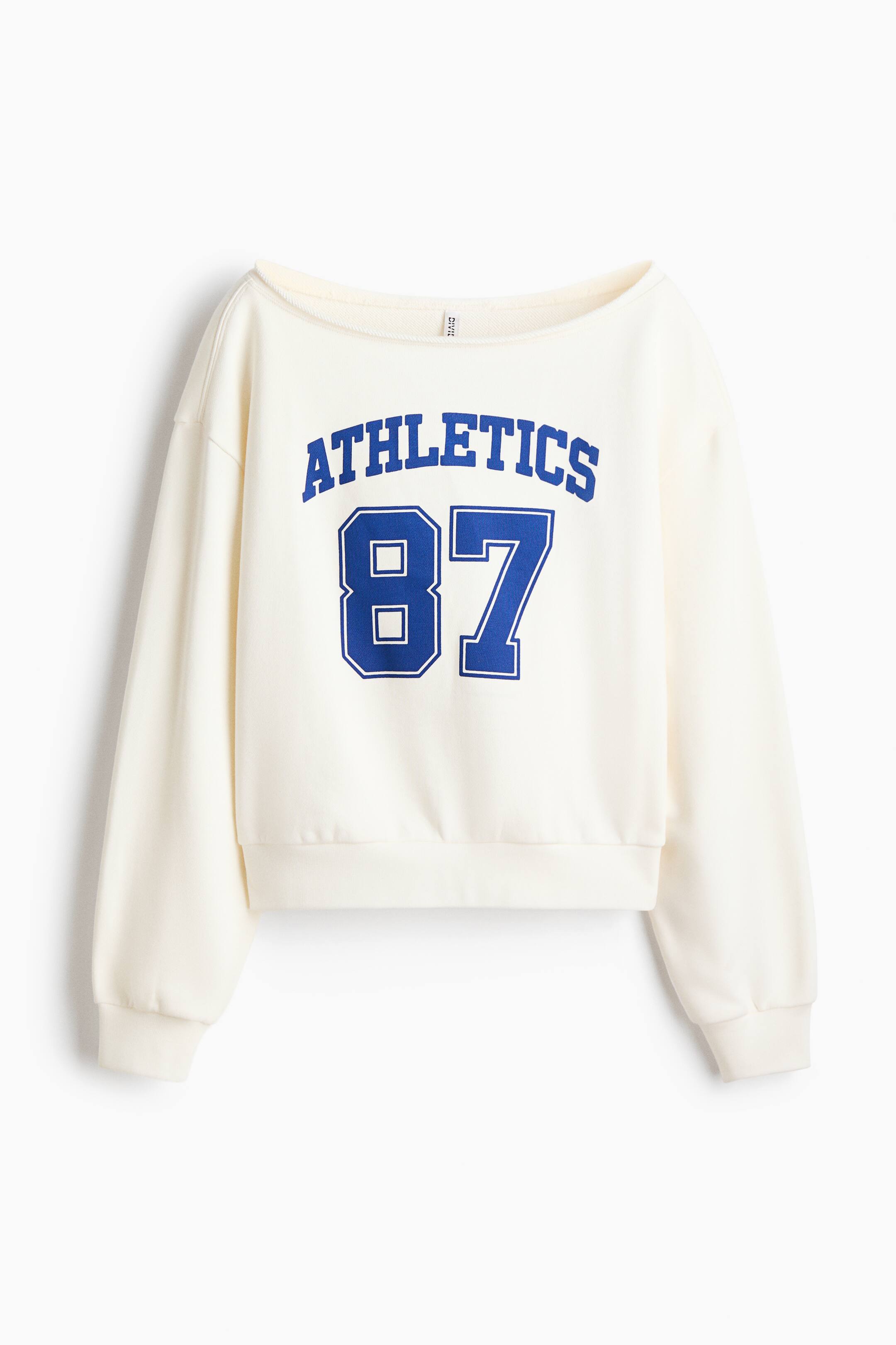 View larger image: A front-facing cream-coloured sweatshirt features a wide boatneck, dropped shoulders, and long sleeves with ribbed cuffs. The chest displays a blue varsity-style graphic with the arched text "ATHLETICS" above the number "87".