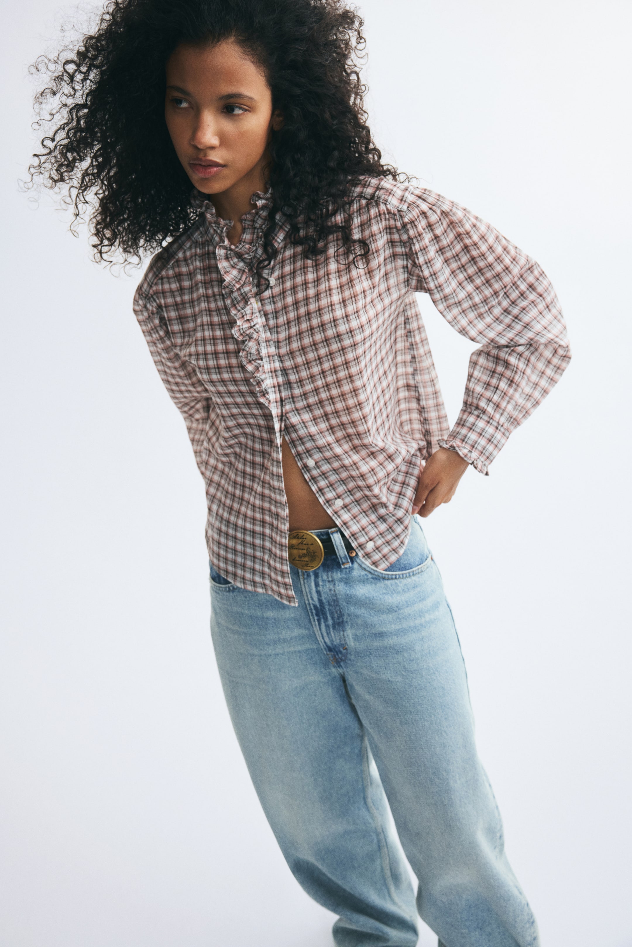Visualizza l'immagine ingrandita: Una blusa a quadri con colletto arricciato, volant frontali abbinati e maniche lunghe è indossata dalla modella. La blusa a quadri marroni e bianchi è abbinata a jeans blu chiaro lavati ed è mostrata parzialmente sbottonata nella parte inferiore davanti mentre la modella posa con una mano sul fianco.
