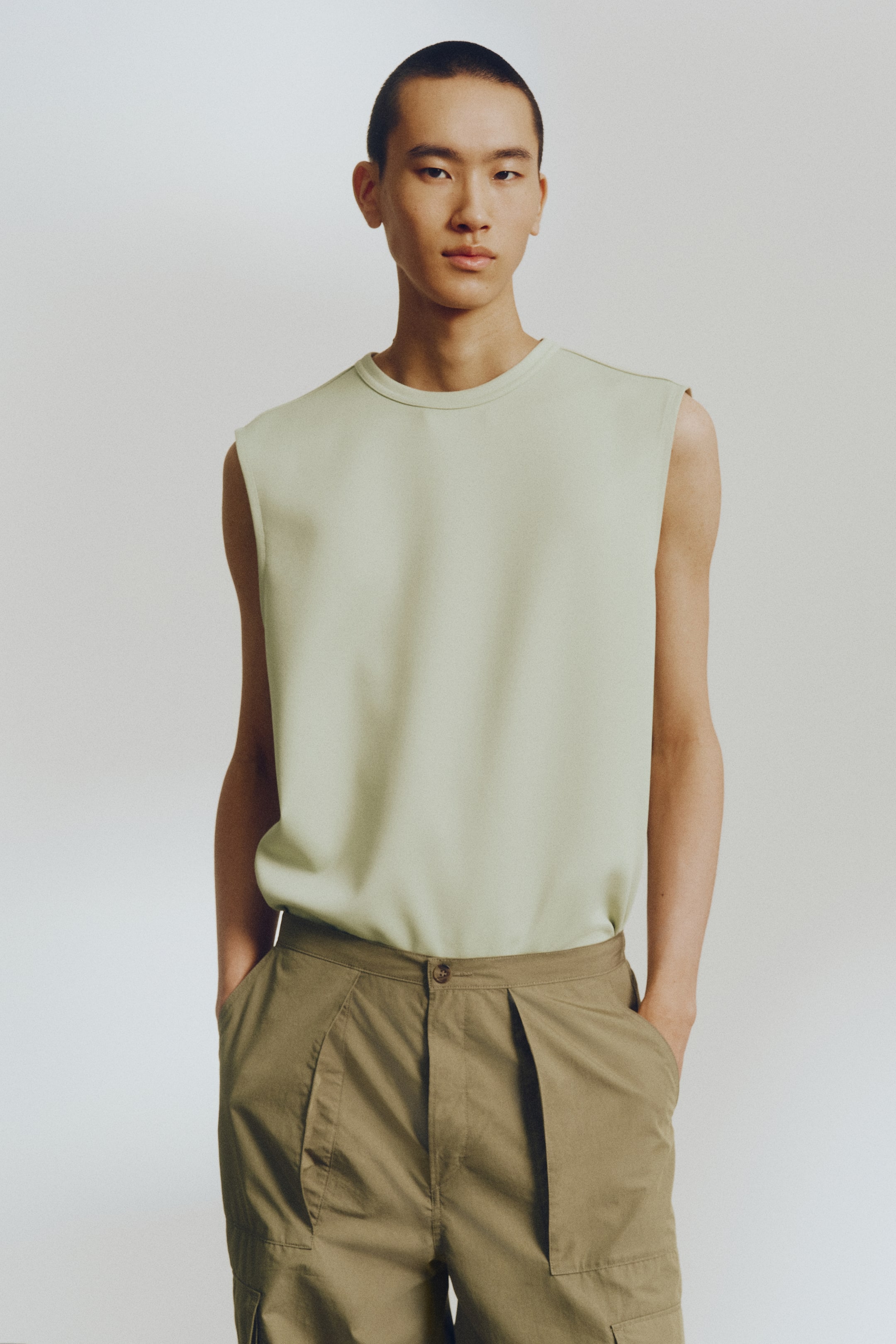 View larger image: A light green sleeveless top is showcased from the front, featuring a crew neck, a relaxed fit, and wide armholes, with its hem tucked into khaki pleated trousers. The trousers feature a front button fastening and two prominent pleats extending from the waistband.