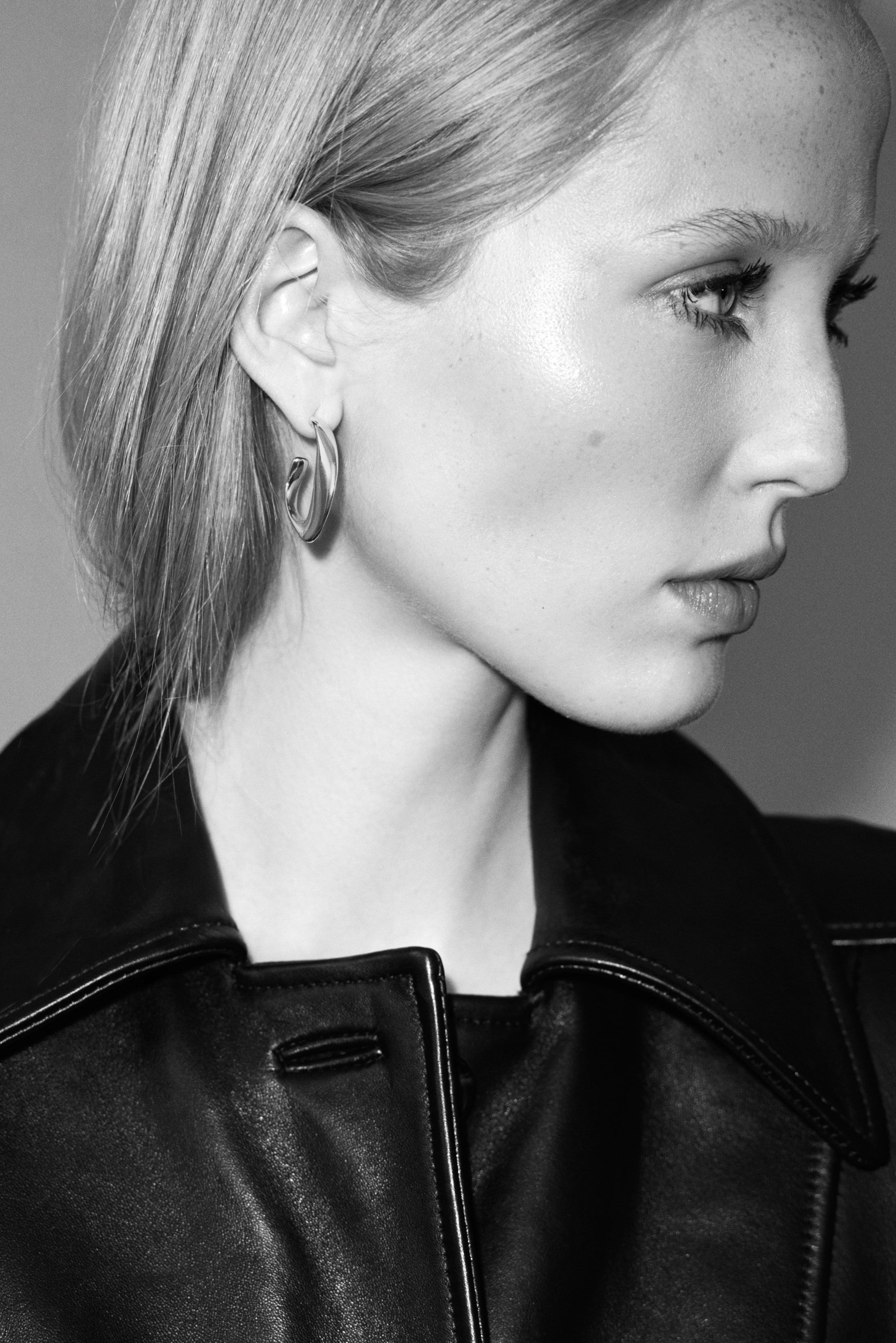Ver imagen más grande: Un pendiente de aro metálico con un diseño robusto y suavemente retorcido se lleva en la oreja de la modelo. El pendiente de aire escultórico se combina con una chaqueta de cuero oscura con el cuello visible en una toma de perfil en primer plano.