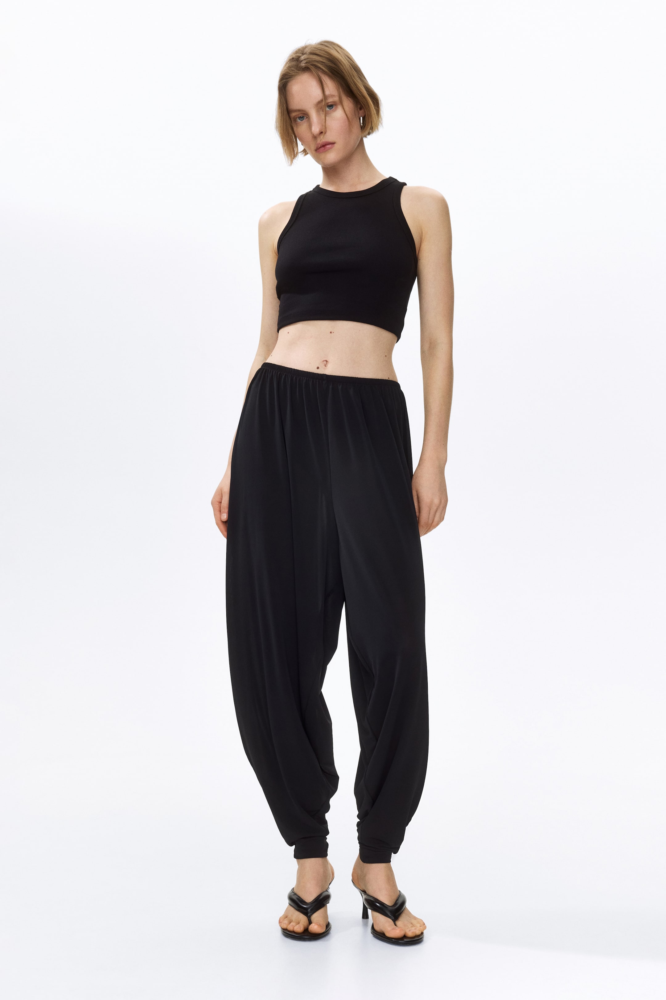Se større billede: Modelen bærer sorte løstsiddende bukser i blød jersey, med elastisk talje og rynker hele vejen rundt, tøndeformede ben og tilspidsede sømme. De er stylet med en sort crop top og sorte hælede sandaler.