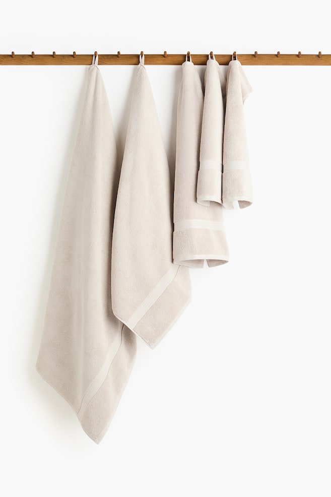 A set of beige cotton terry towels in varying sizes is hanging, each towel featuring a subtle sheen, woven trim details along its short sides, and a visible loop for hanging. - 1