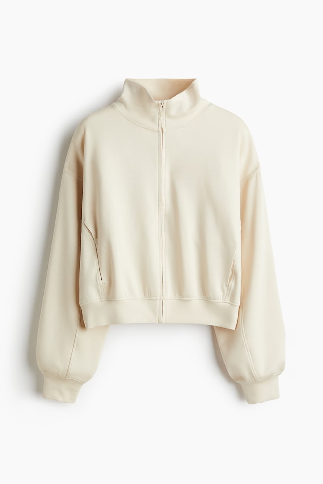 A cream-coloured zip-up jacket is being displayed from the front, featuring a high stand-up collar, a full-length front zipper, and concealed side pockets. It is showcasing dropped shoulders, voluminous sleeves, and ribbed cuffs and hem. - 1