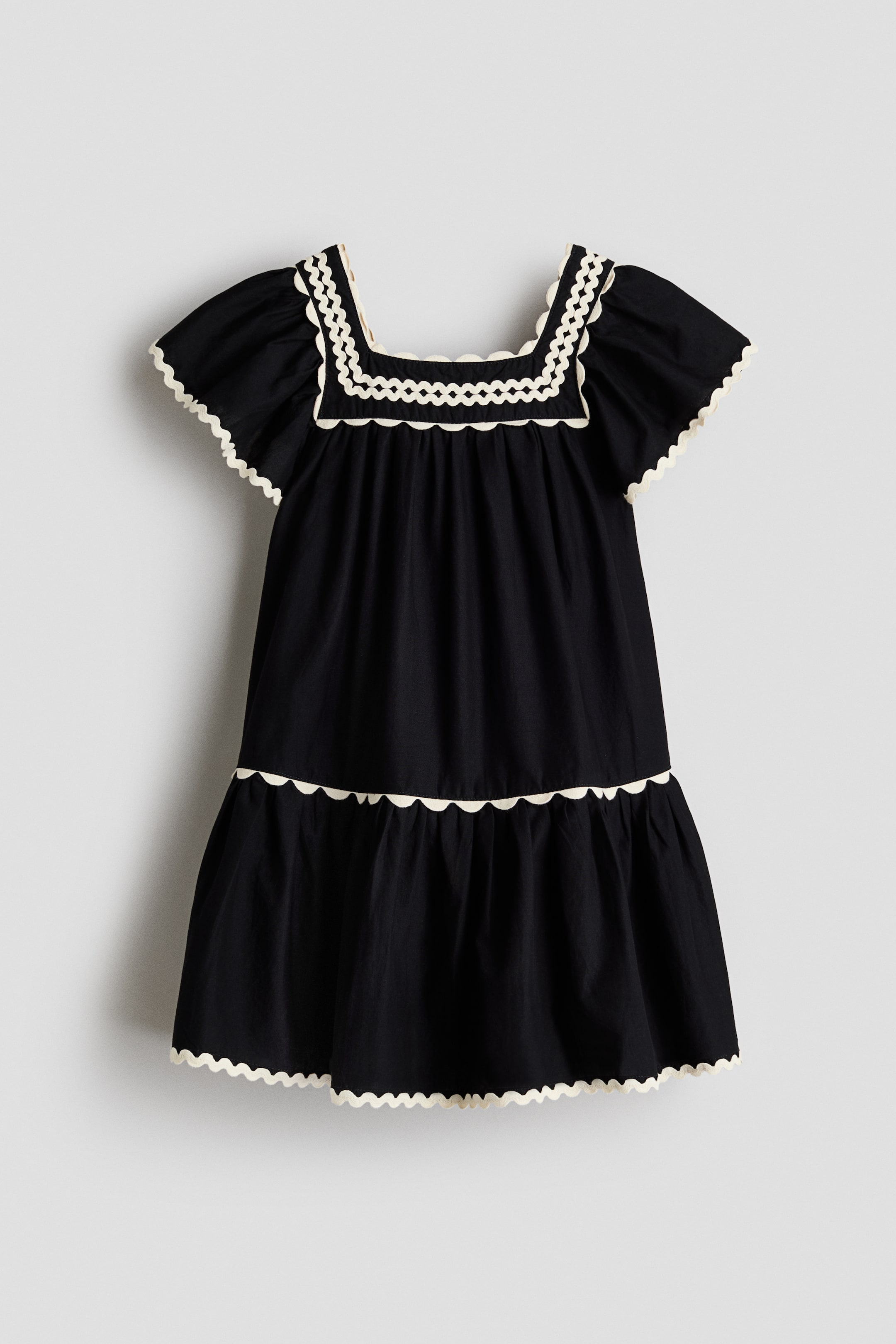 Größeres Bild ansehen: Ein schwarzes ausgestelltes Kleid wird von vorne gezeigt, mit einem quadratischen Ausschnitt und Schmetterlingsärmeln. Es ist mit kontrastierenden cremefarbenen Muschelkanten entlang des Ausschnitts, der Ärmel und des Stufenrocks verziert, wobei die Ausschnittverzierung auch ein inneres Wellenmuster aufweist.